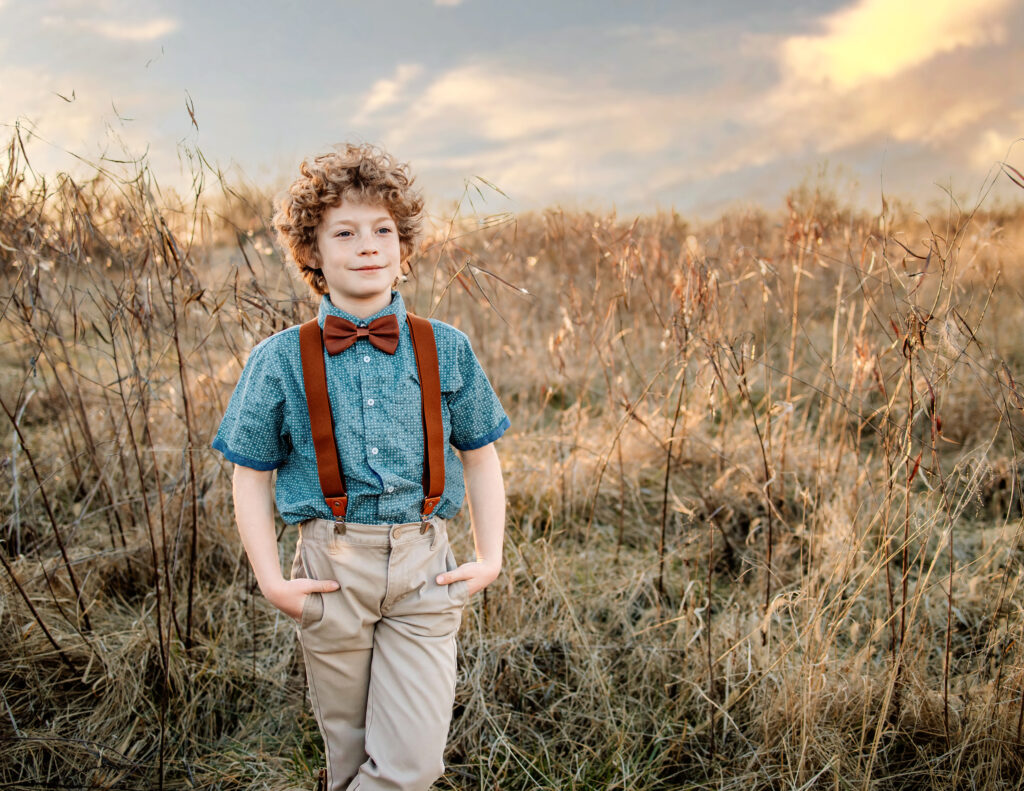 A handsome young boy posed in a field of tall grass with the sunset behind him.