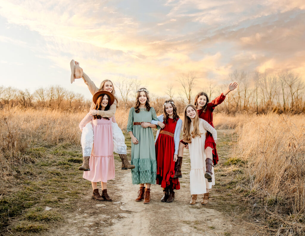 6 girls walking on a dirt road wearing beautiful dresses, some are piggy back, and having fun.