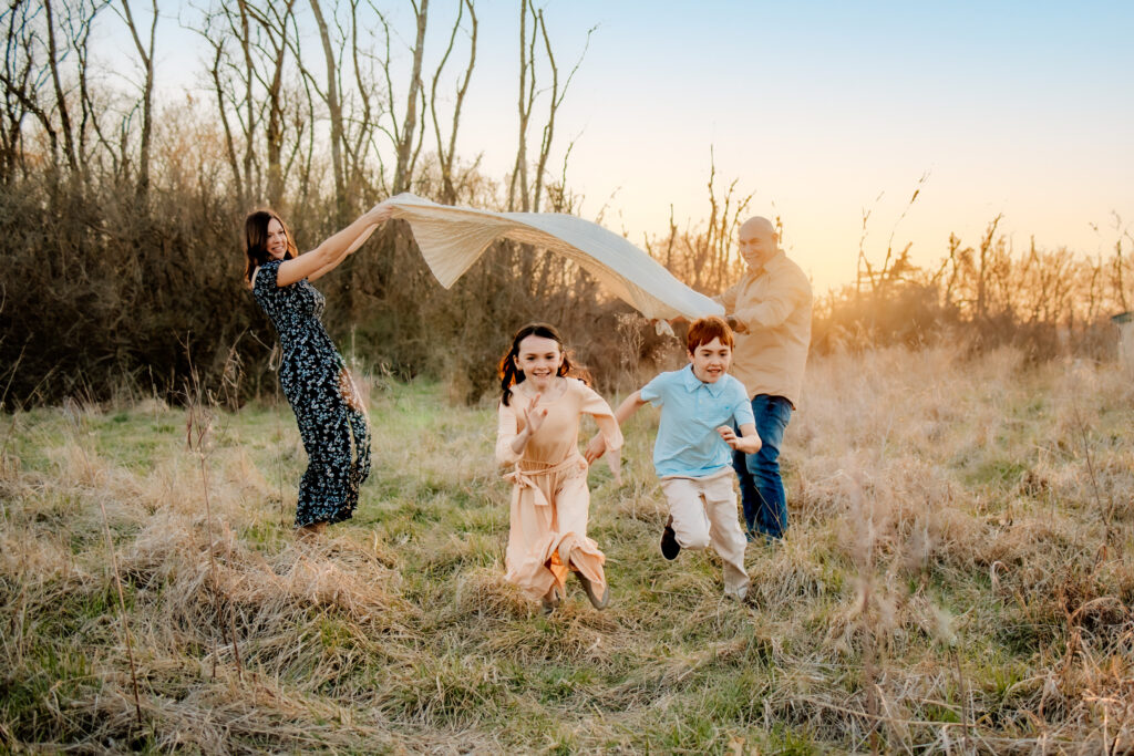 a little boy and a little girl are running under a white blanket that their mom and dad are fluffing into the air.