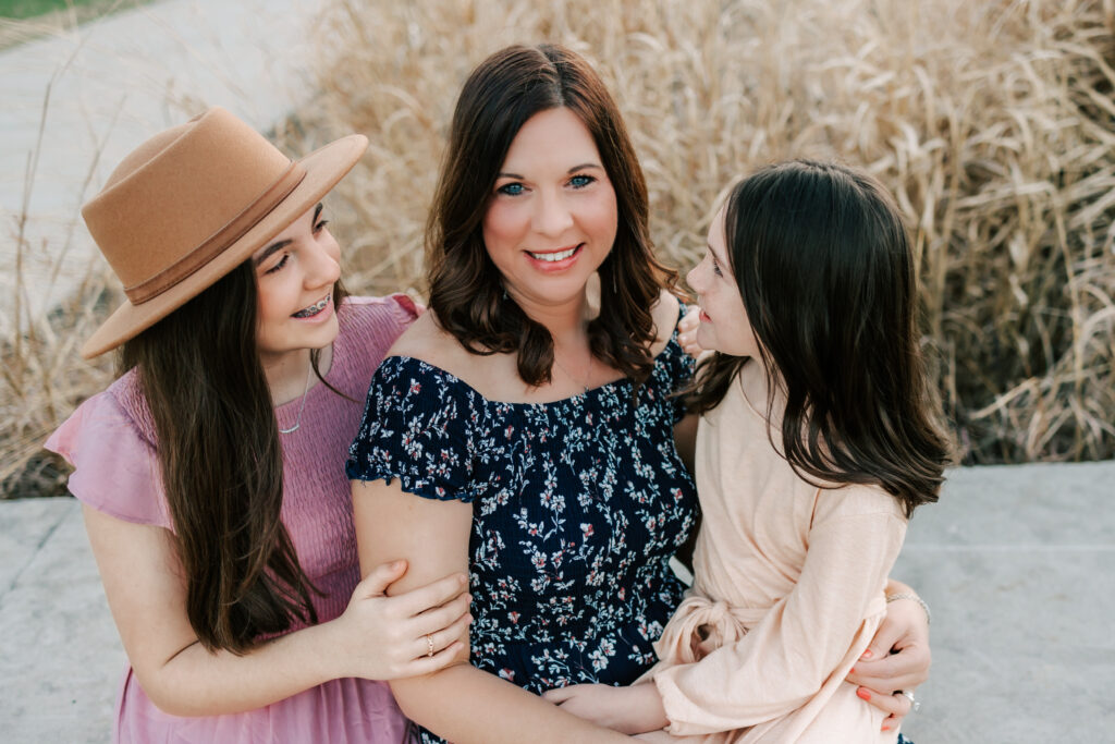 A mother sitting on a ledge with her two daughters, one on either side of her. Mom is looking at the camera and the two girls are looking at their mom.