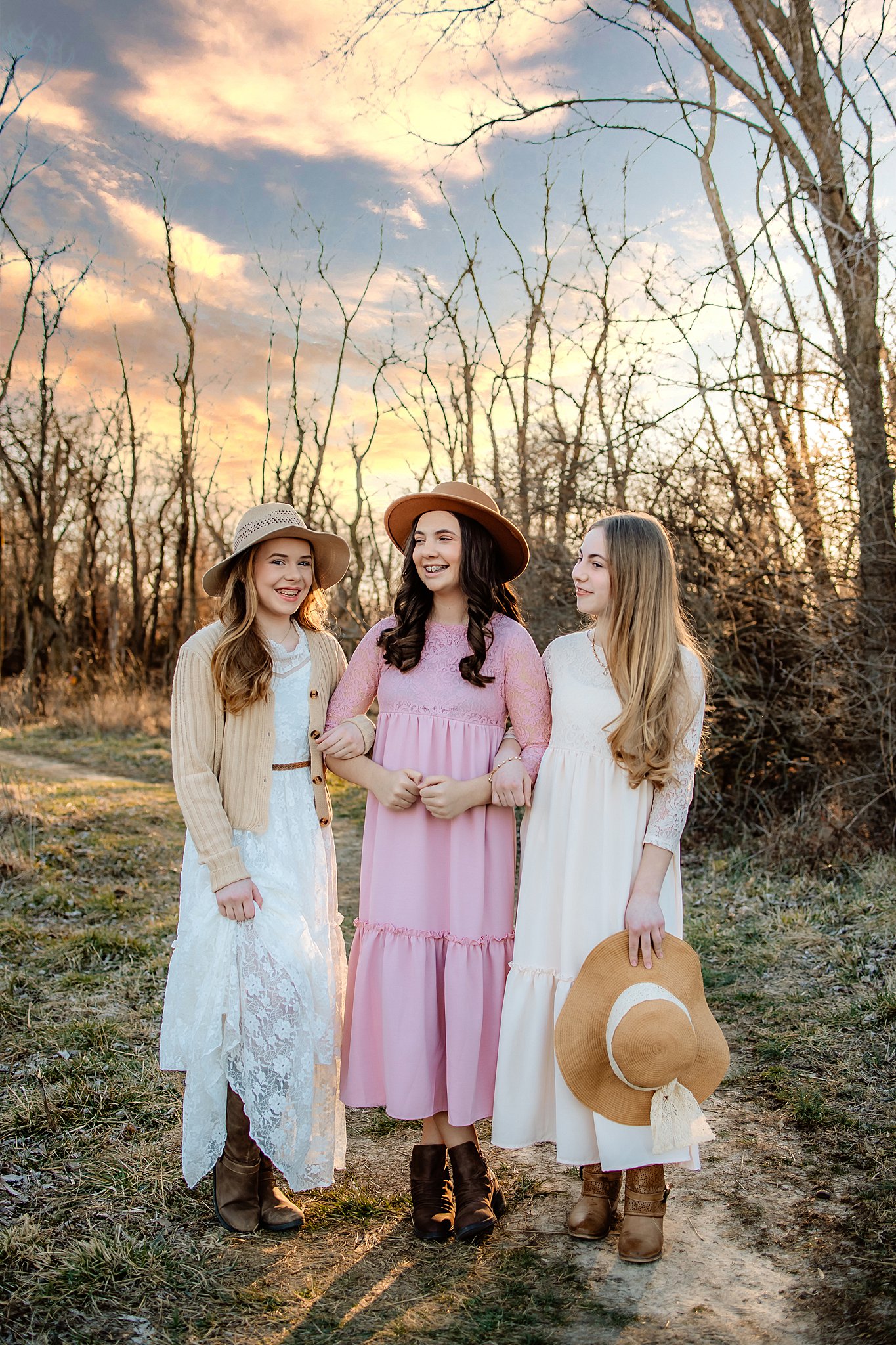 Three young girls stand arm in arm in a dirt path at sunset in long dresses and woven hats