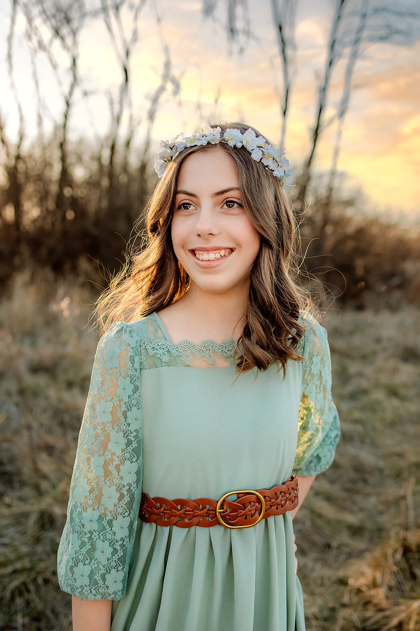 A girl with long curly brown hair stands in a field at sunset in a green dress with lace sleeves and a flower headband hair salons in saint louis