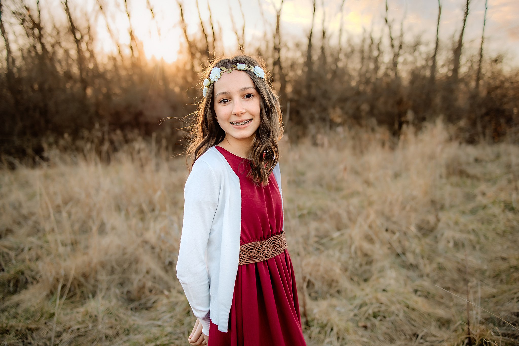 A young girl in a red dress and white sweater stands in a field of golden grass with hands behind her back