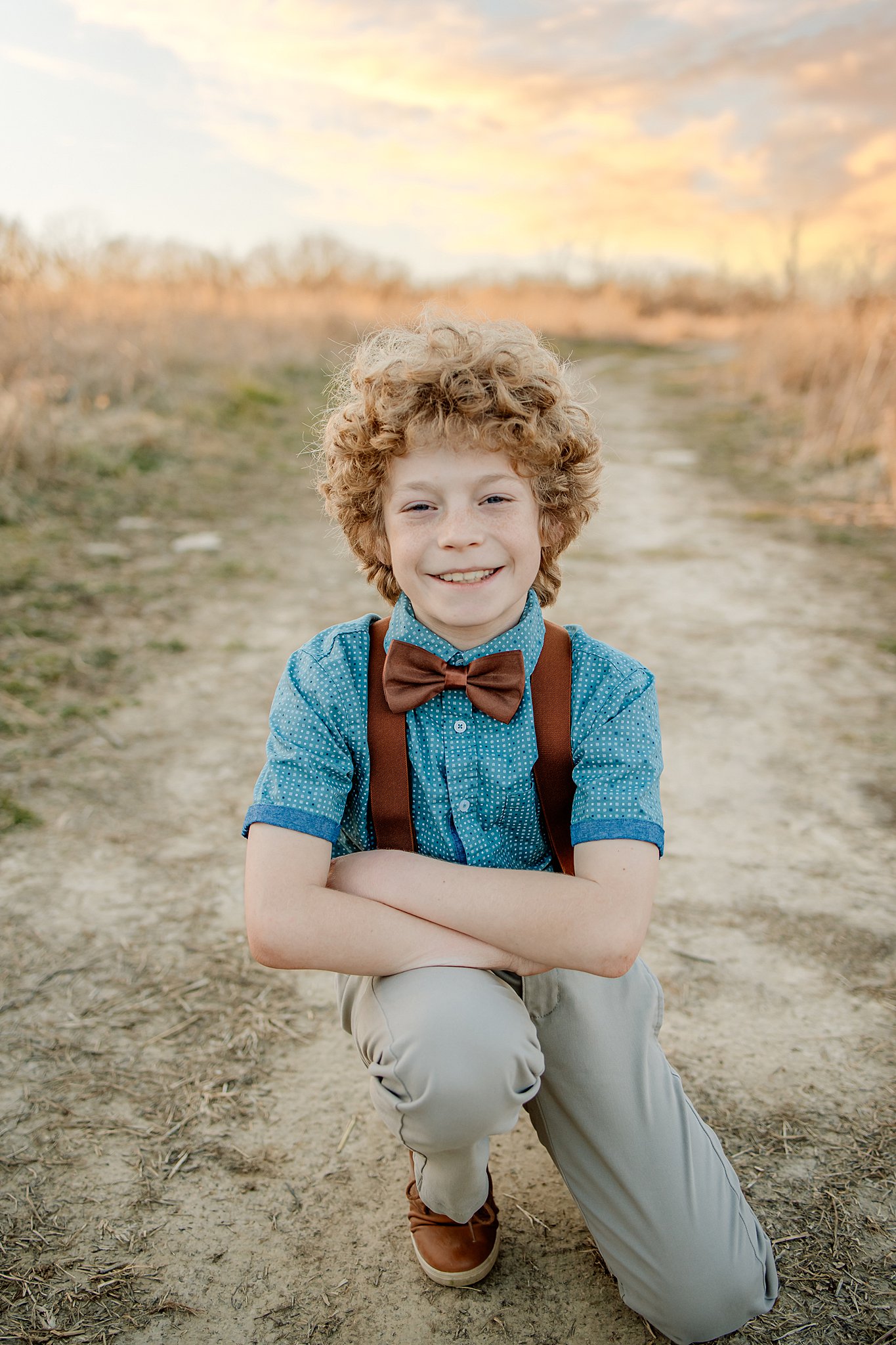 A young boy kneels in khaki pants, blue shirt and brown bowtie and suspenders in a dirt path oakville pediatricians