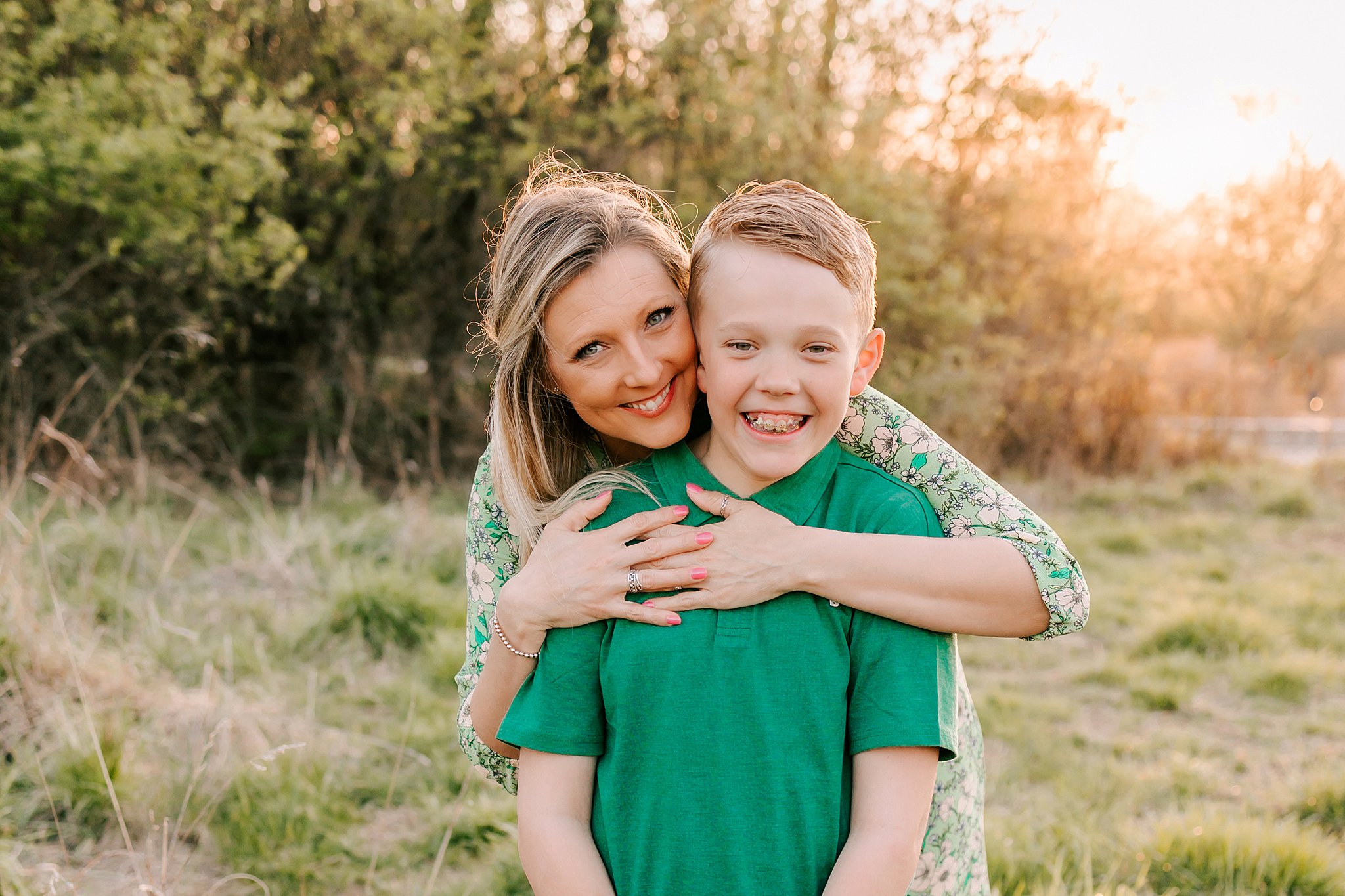 A mother in a green floral dress leans over the shoulder and hugging her son in a green polo while in a park at sunset spas in oakville