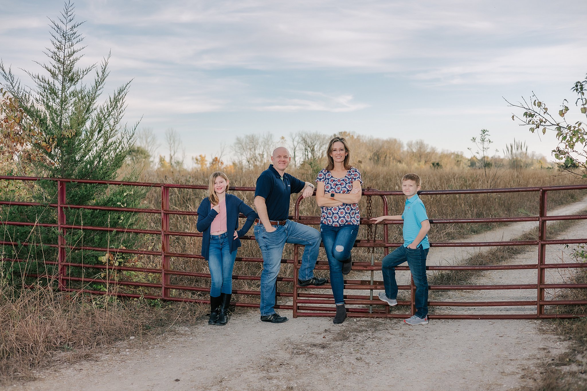 A family of four leans against a farm gate on a dirt road in front of a field of tall grass