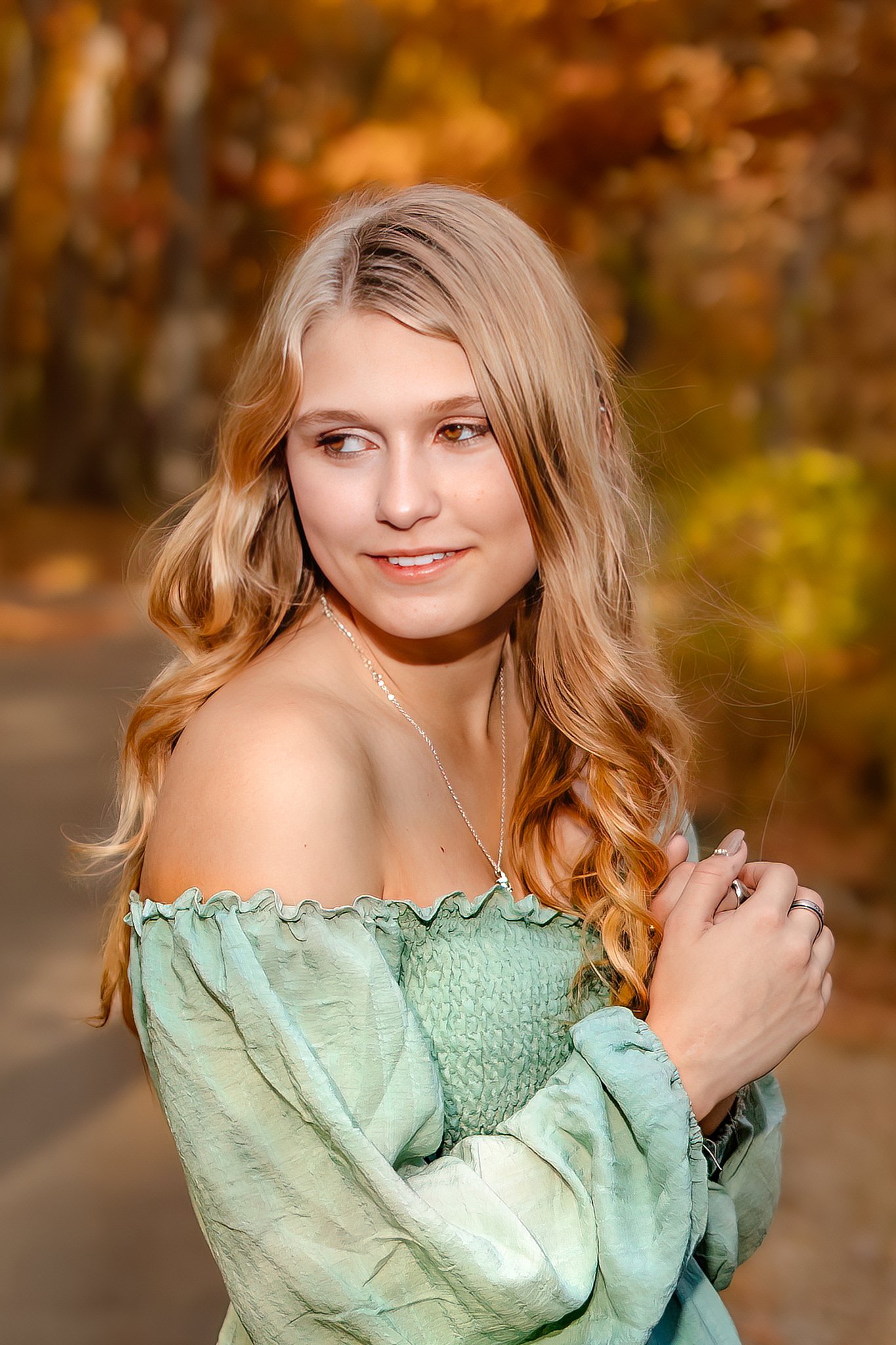 A high school senior in a green dress stands in a park path looking over her shoulder