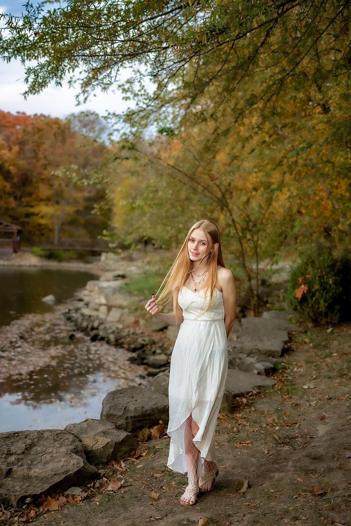 A high school senior in a white dress walks along a park pond playing with her hair boutiques in St Louis