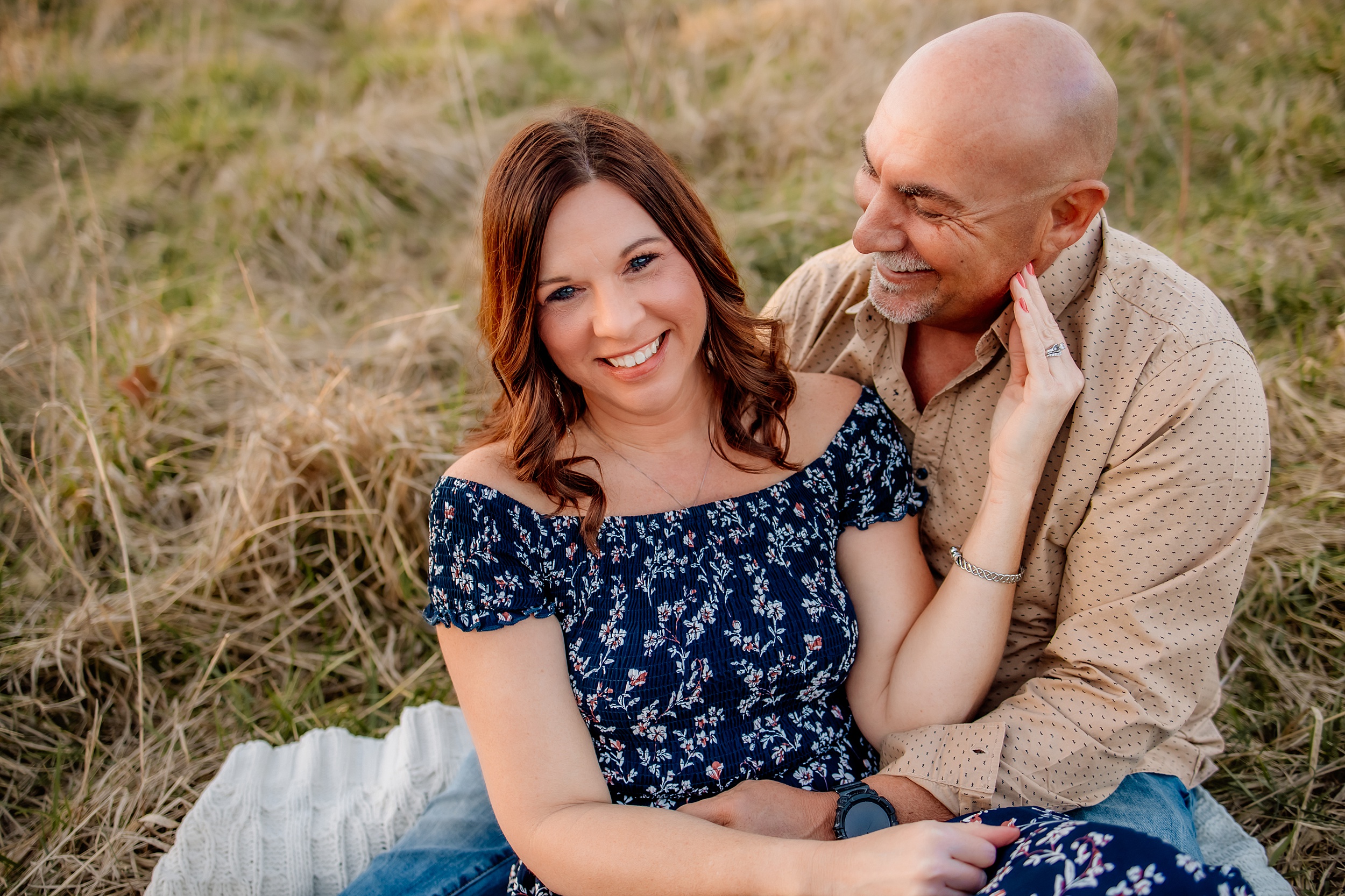 A husband and wife cuddle and laugh together on a picnic blanket in a field of grass