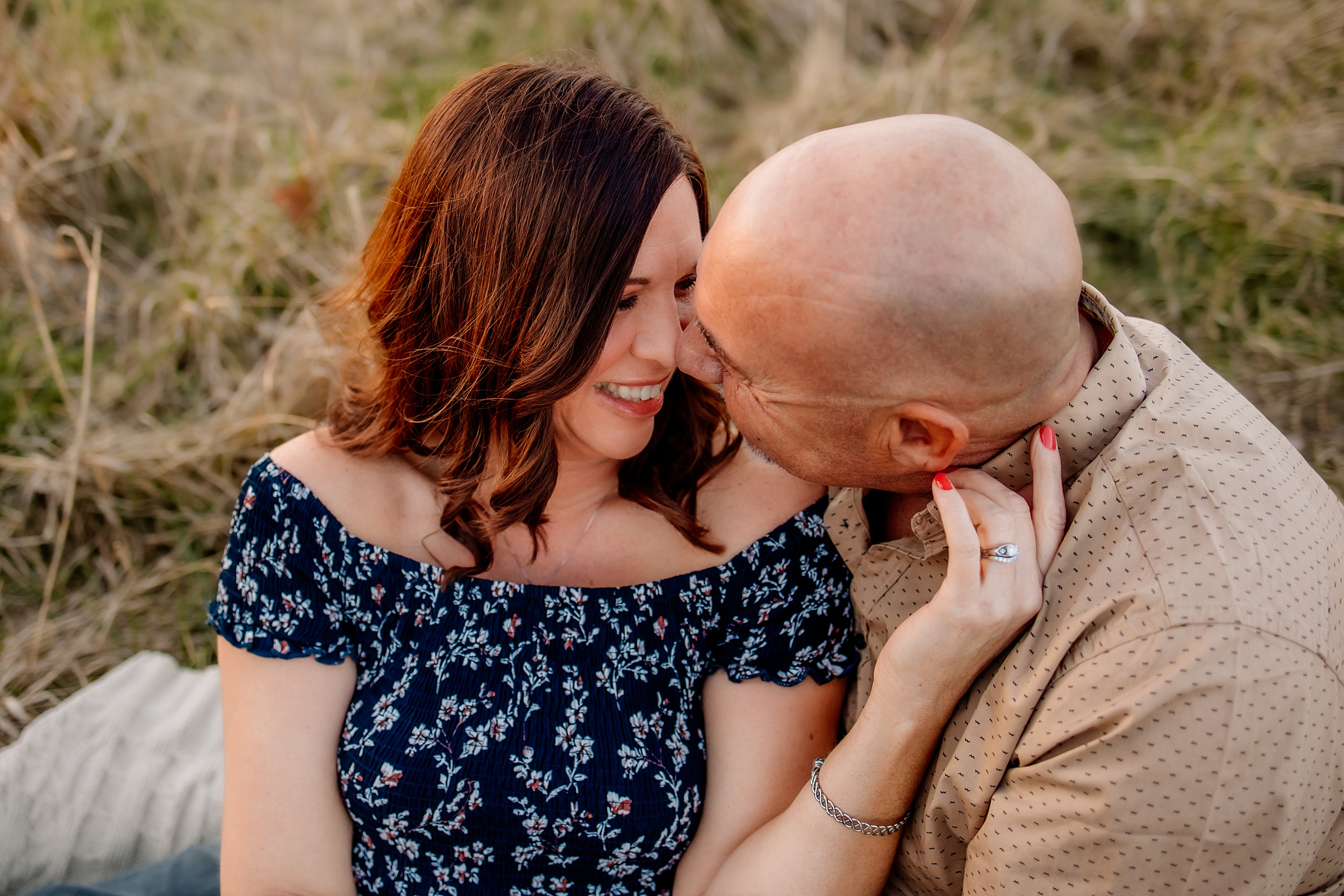 A husband and wife sit on a picnic blanket at sunder while the man leans in for a kiss spas in st louis