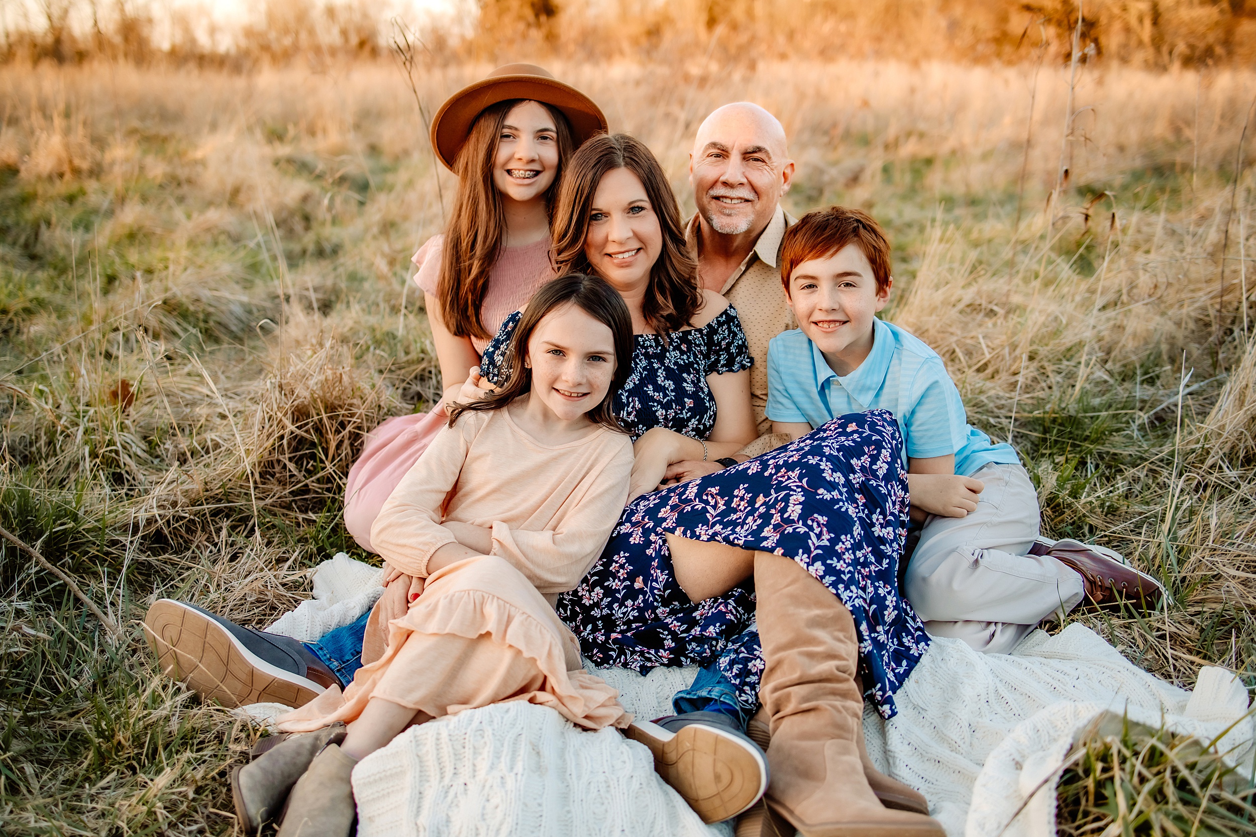 A mother and father sit on a blanket in a fieeld of tall grrass with their three kids cuddling in with them spas in st louis