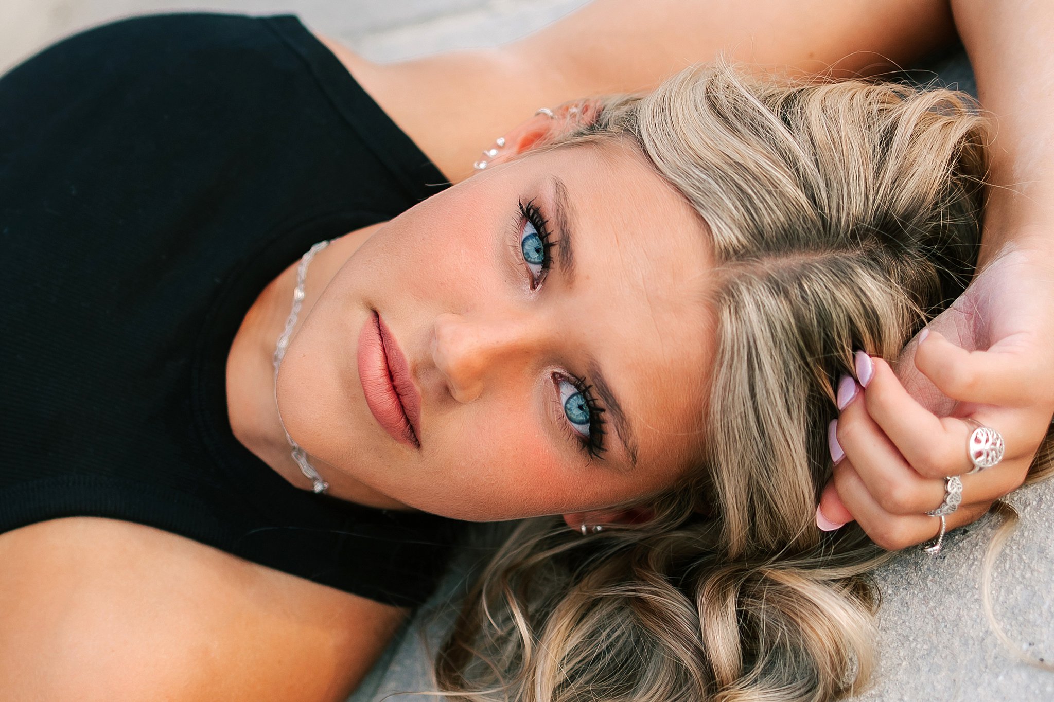 A high school senior lys on some concrete in a black blouse and lots of jewelry