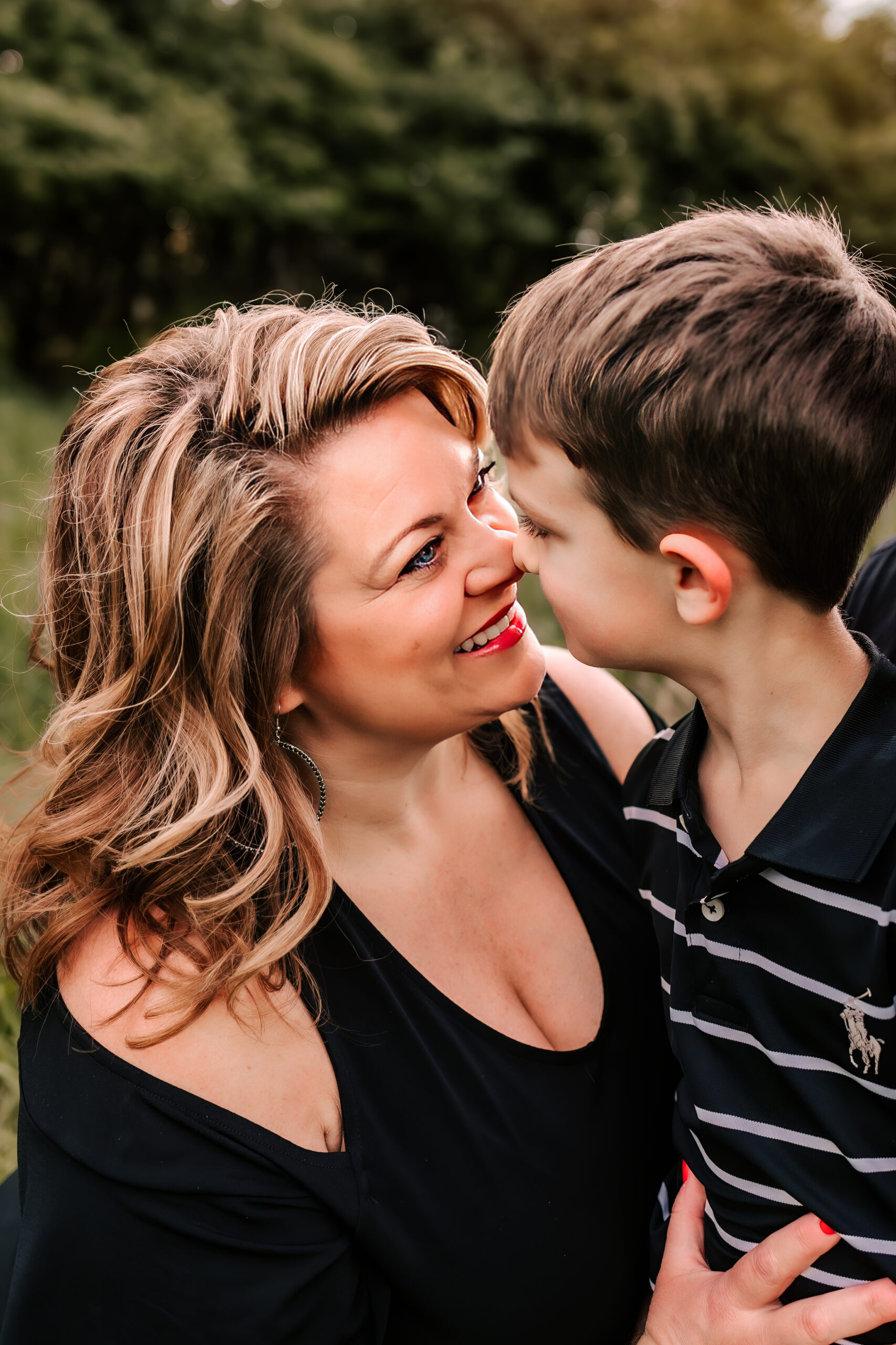 A mother presses her nose against her young son's nose while sitting in a field at sunset