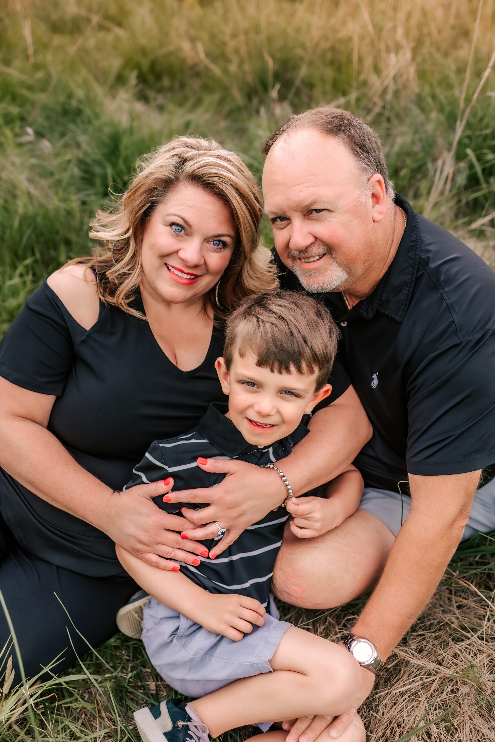 A mother and father in black sit in a field of tall grass hugging their young son between them st louis pediatricians