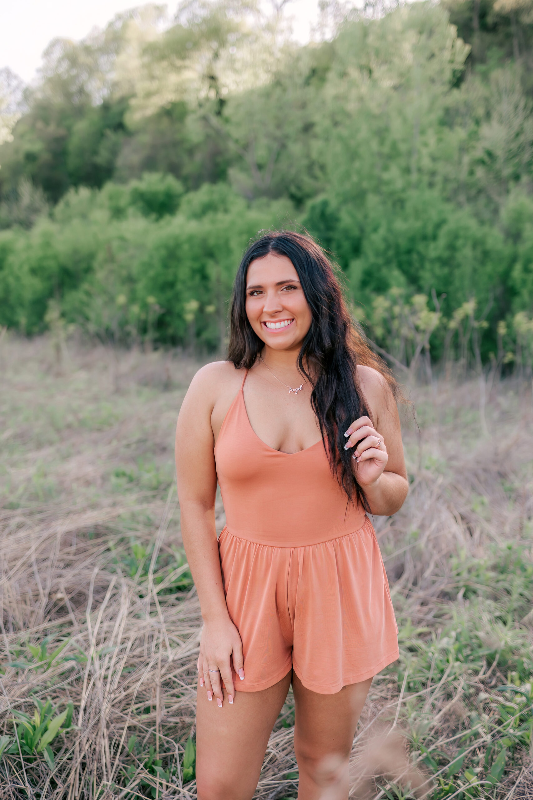 A high school senior stands in a field of tall grass with a hand in her hair in aa pink dress