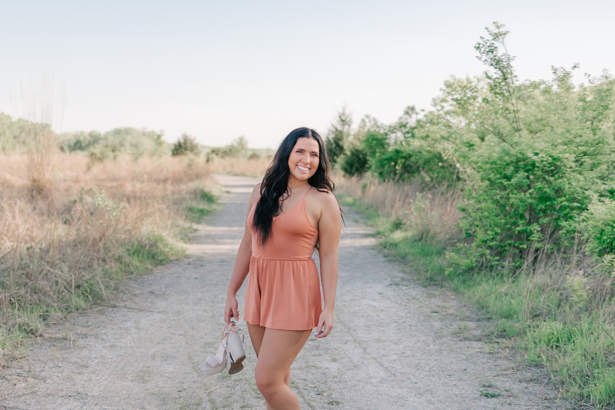 A high school senior stands in a gravel park path in a pink dress holding her shoes in her hand st louis tutors