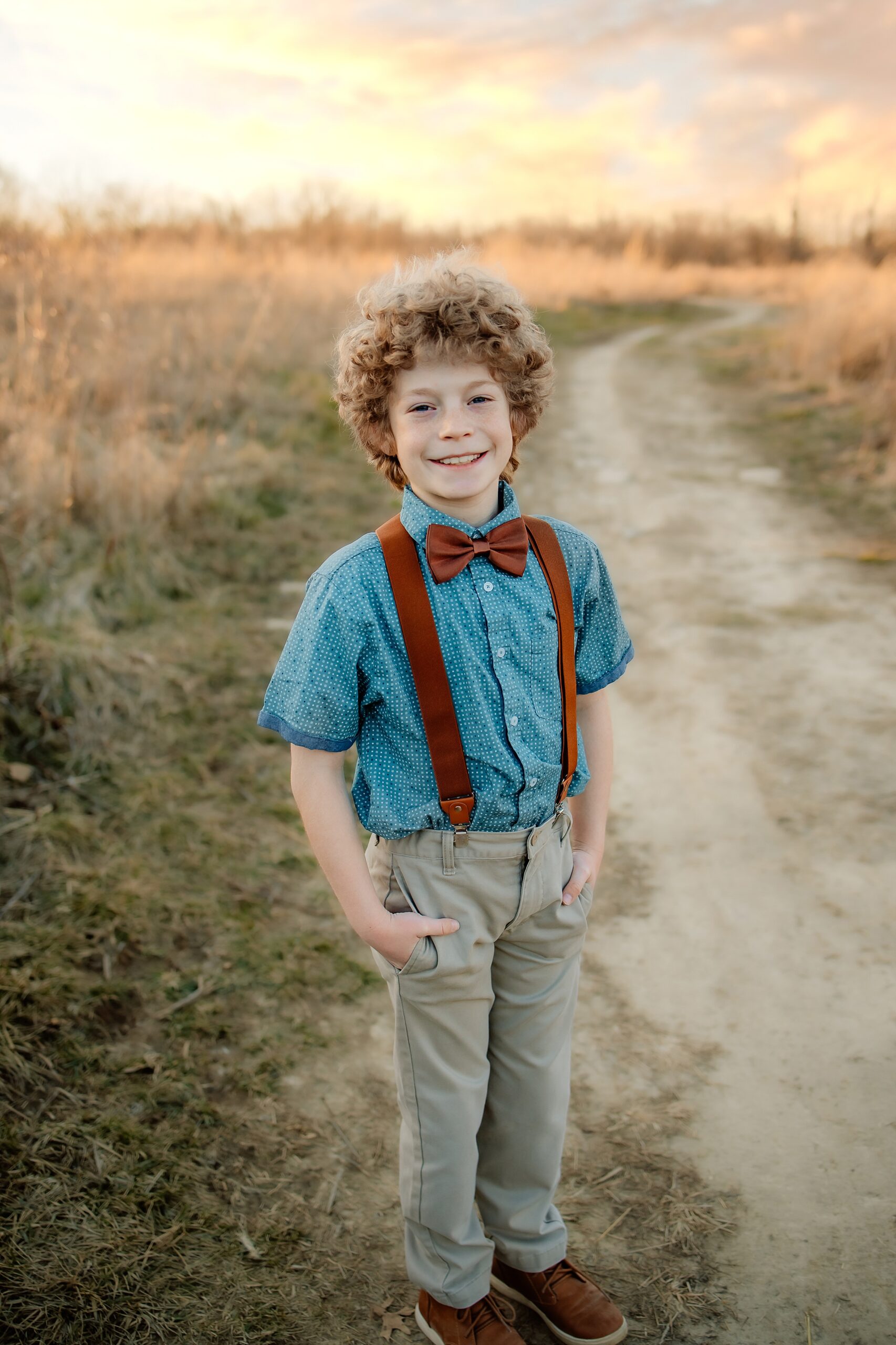 A young boy in khaki pants, suspenders and a bowtie stands in a dirt trail through tall grass at sunset