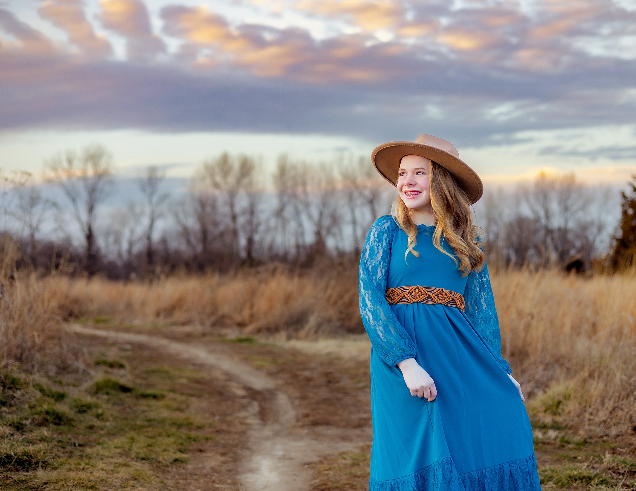 A high school senior twirls her blue dress while walking through a park trail in a large hat st louis dance studios