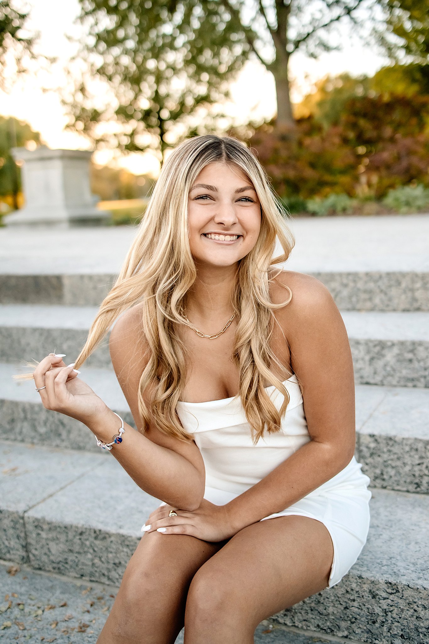 A high school senior in a white dress sits on steps in a park twirling her hair