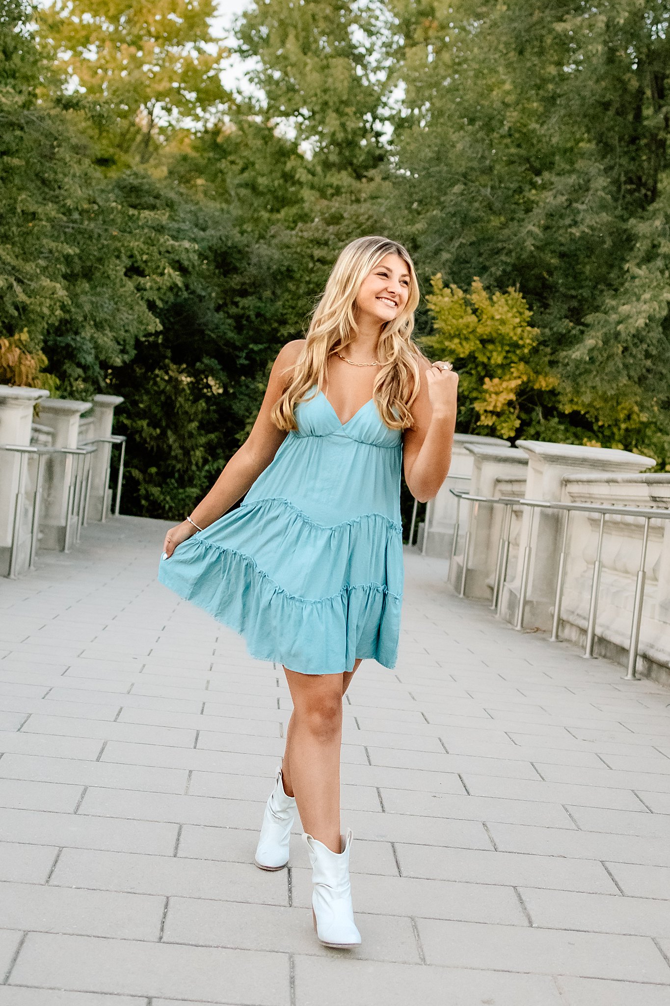 A high school senior in a blue sun dress walks across a bridge in white boots st louis nail salons