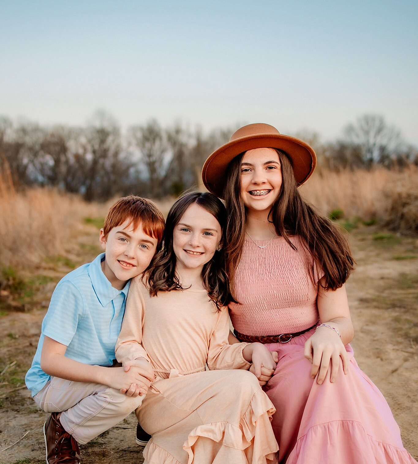 Two sisters in long dresses sit in a park trail holding hands with their brother in a polo and khakis st louis pediatric dentist
