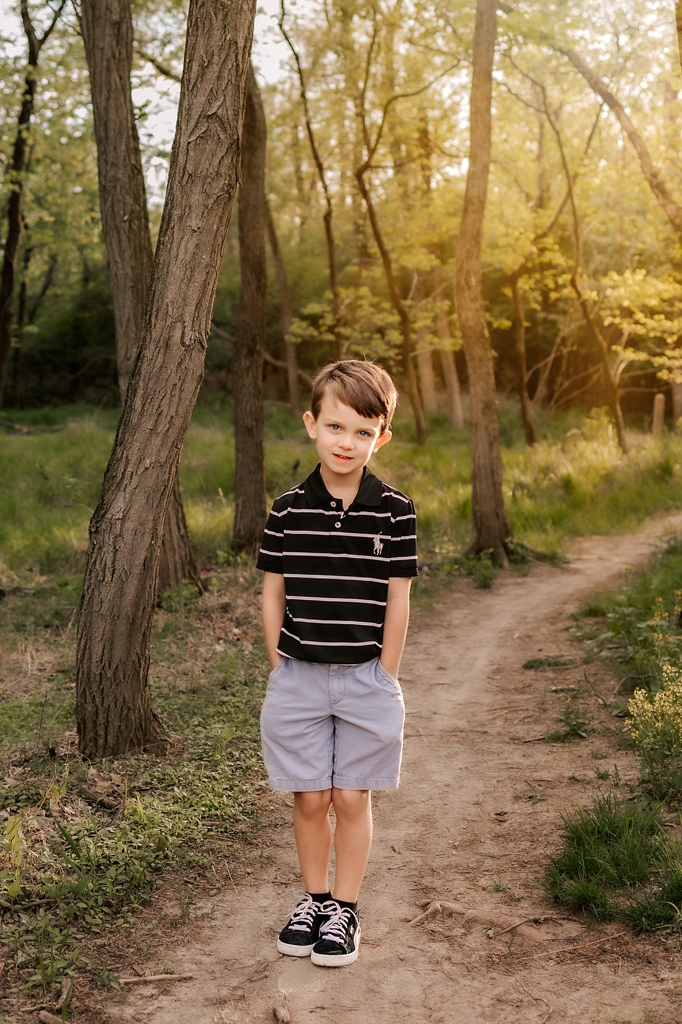 A young boy in a striped polo shirt stands in a wooded trail with hands in his pockets st louis preschools