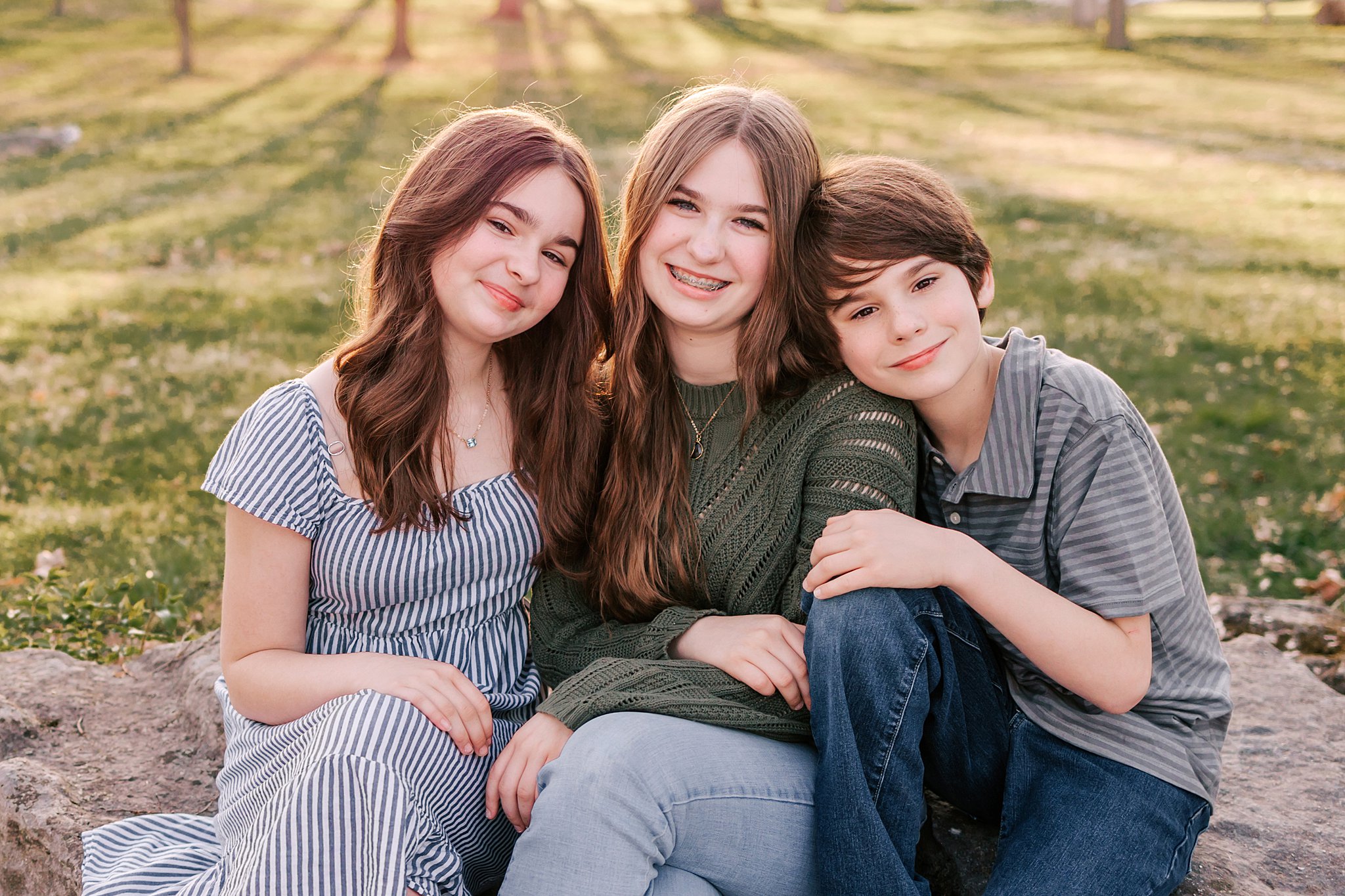 Three siblings sit together on a large rock in a park at sunset