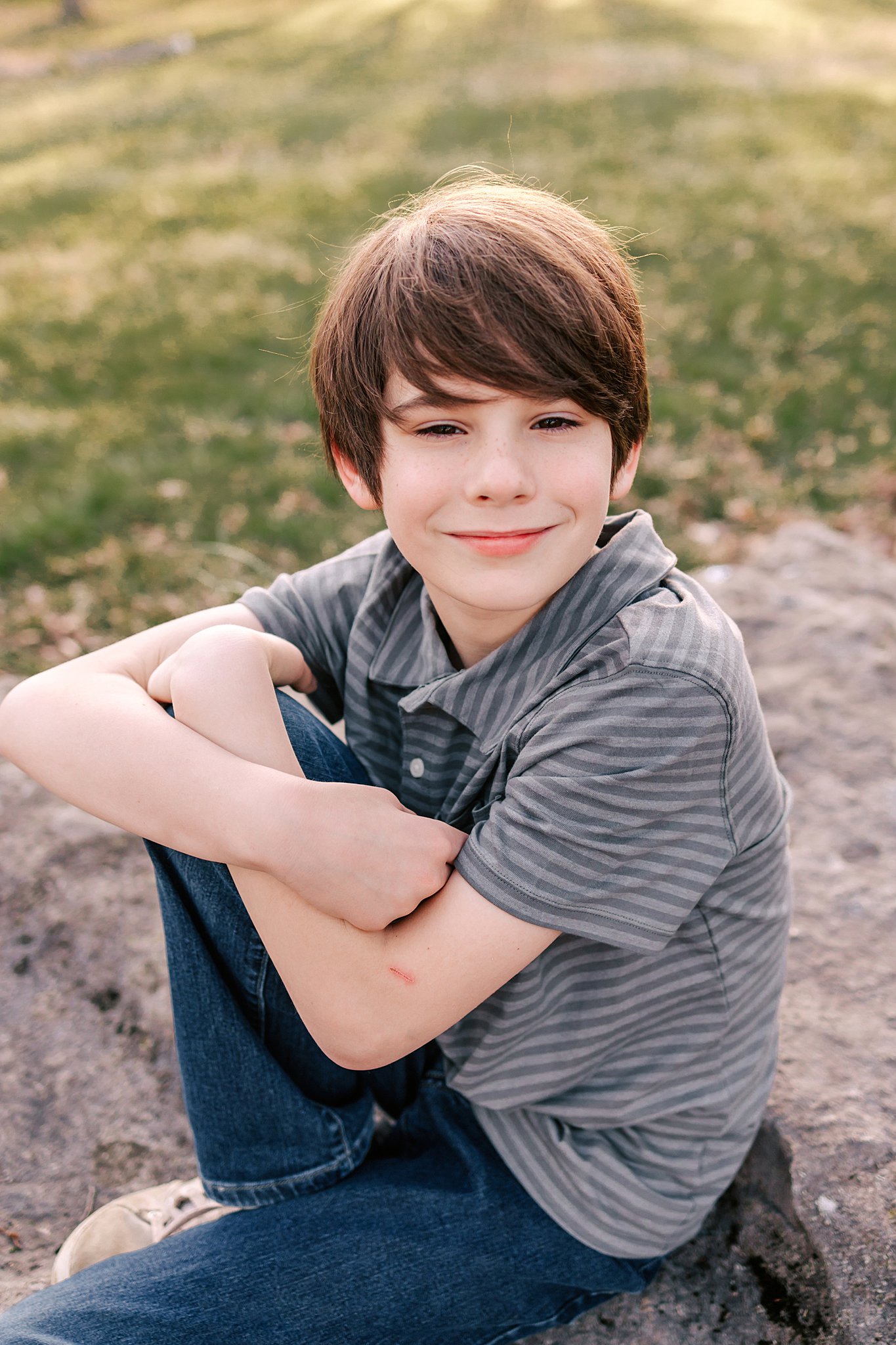 A young boy in a stripe polo shirt sits on a rock in a park st louis youth soccer clubs