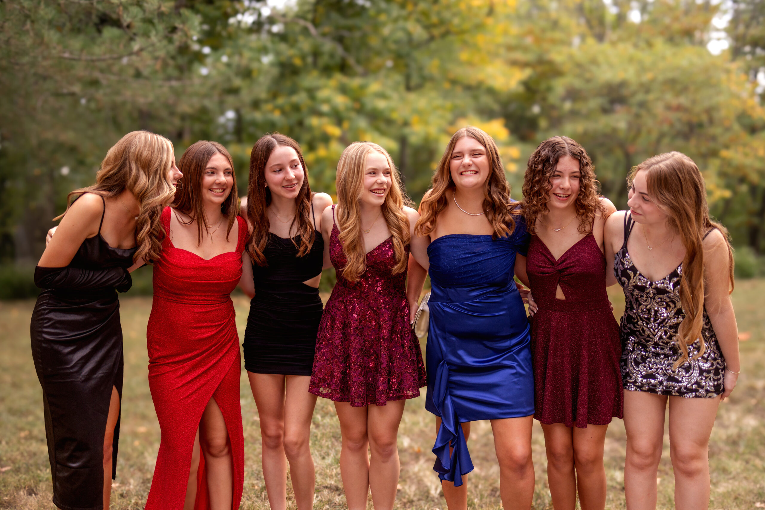 A group of 7 girls are walking in a line with their arms around each other and looking and laughing with each other. They are wearing prom dresses and preparing for the ultimate prom guide.