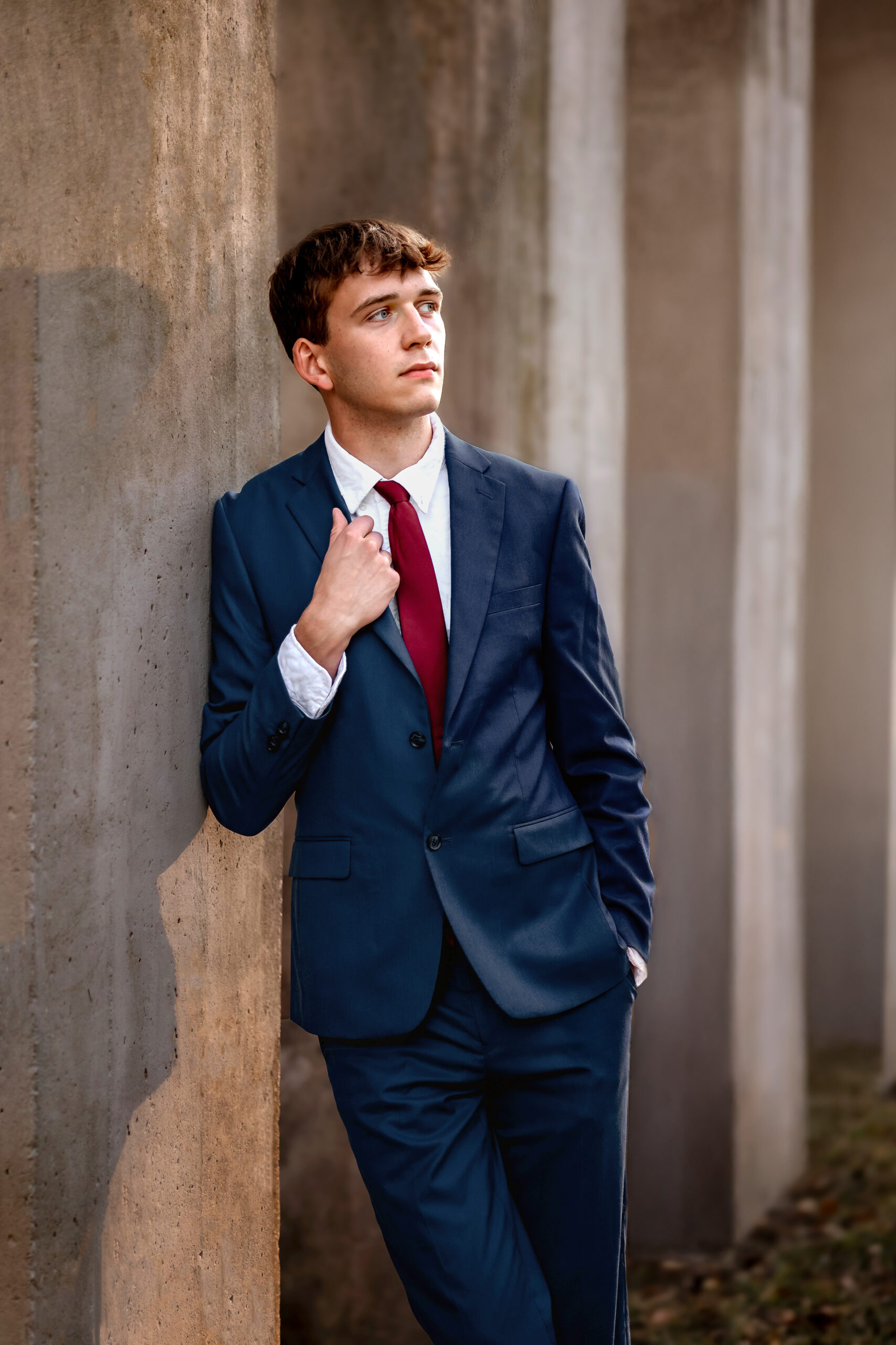 A young man is dressed in a blue suit with a red tie. He's leaning against a pillar with his hand on the jacket lapel posing for his prom photos.