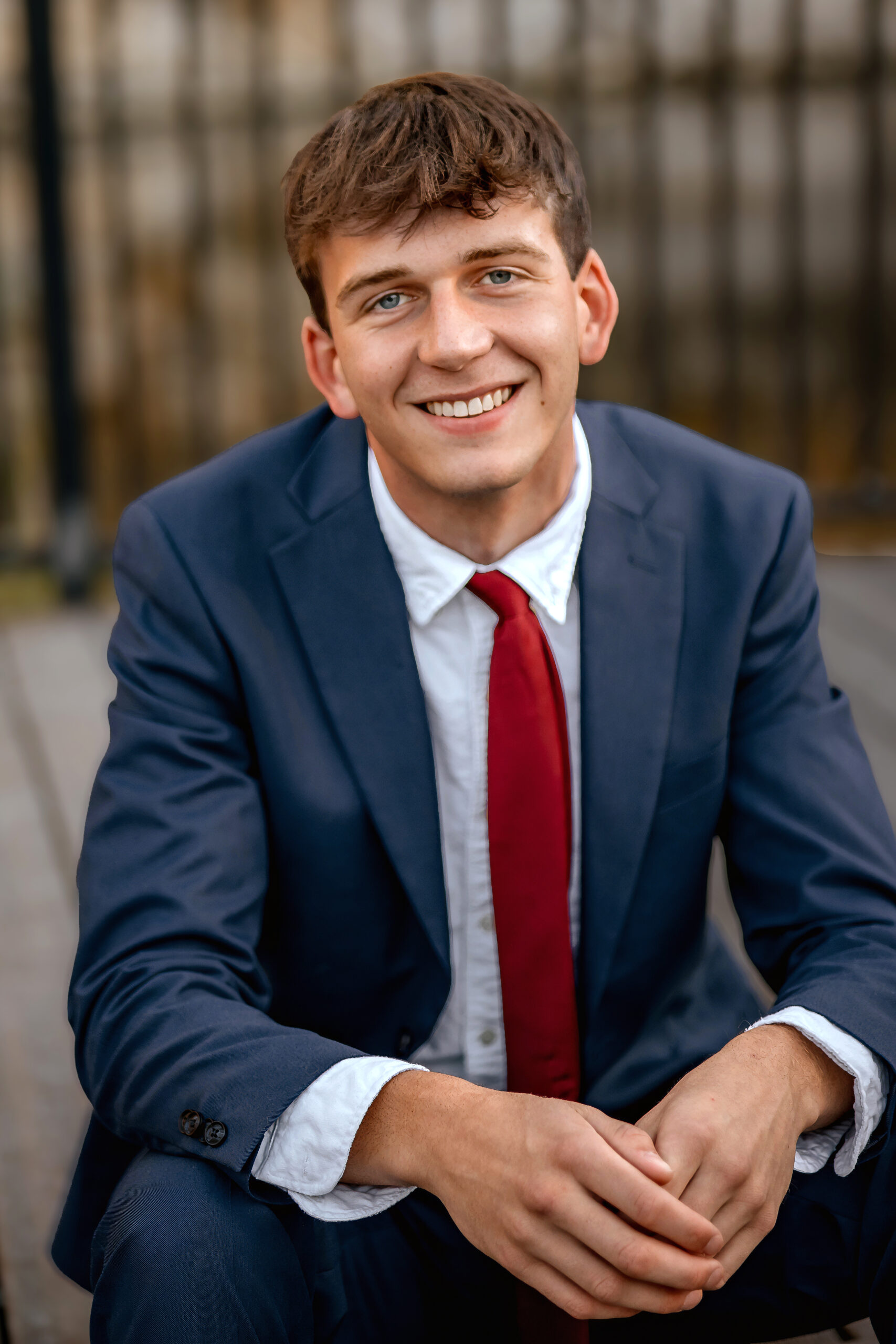 A close up image of a young man in a blue suit with a red tie. He is sitting on a step at a park in St. Louis with his elbows resting on his knees posing for his Prom photo session.