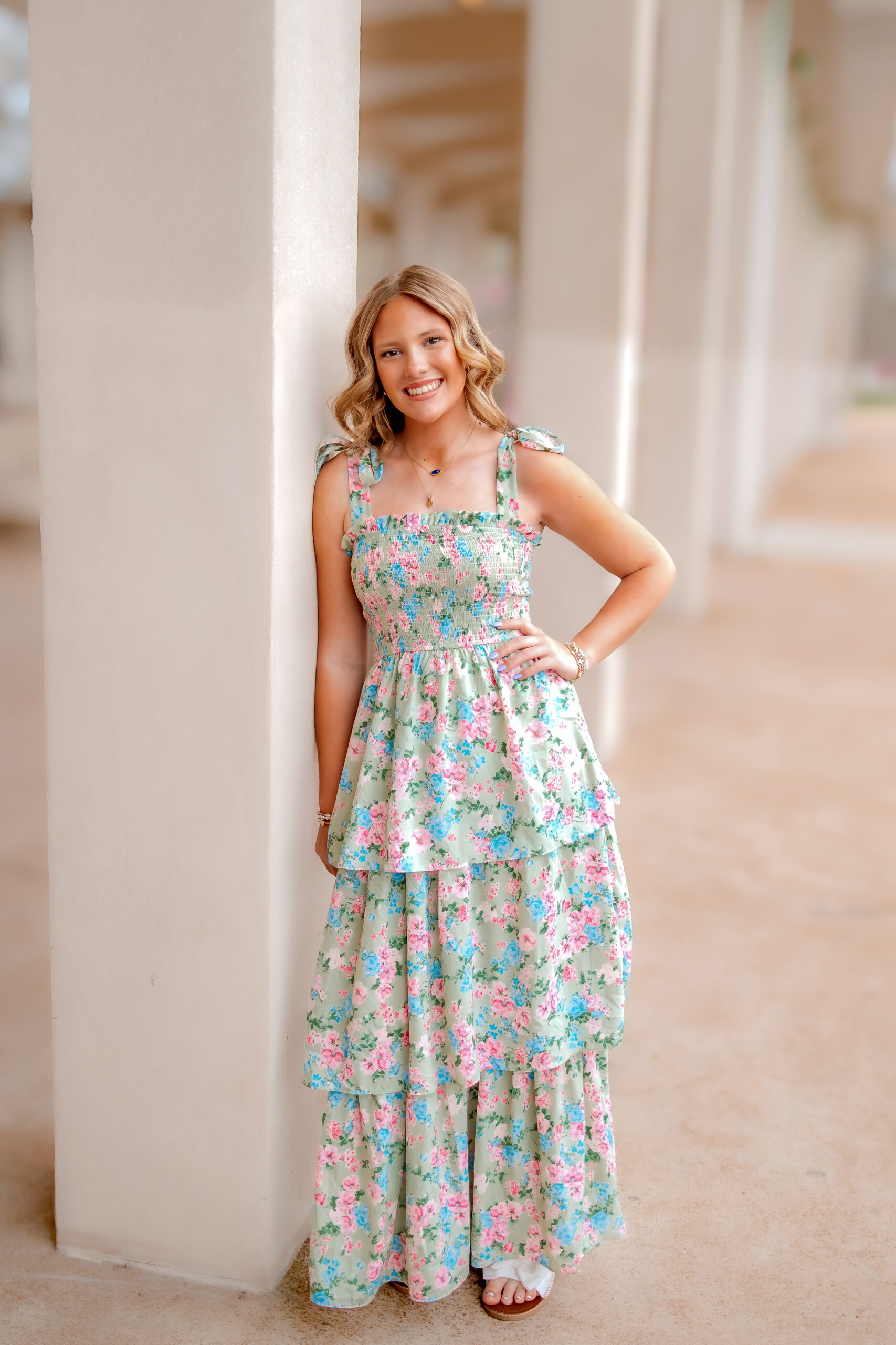 A girl in a ruffled long floral dress is leaning against a pillar with her left hand on her hip. she is smiling.