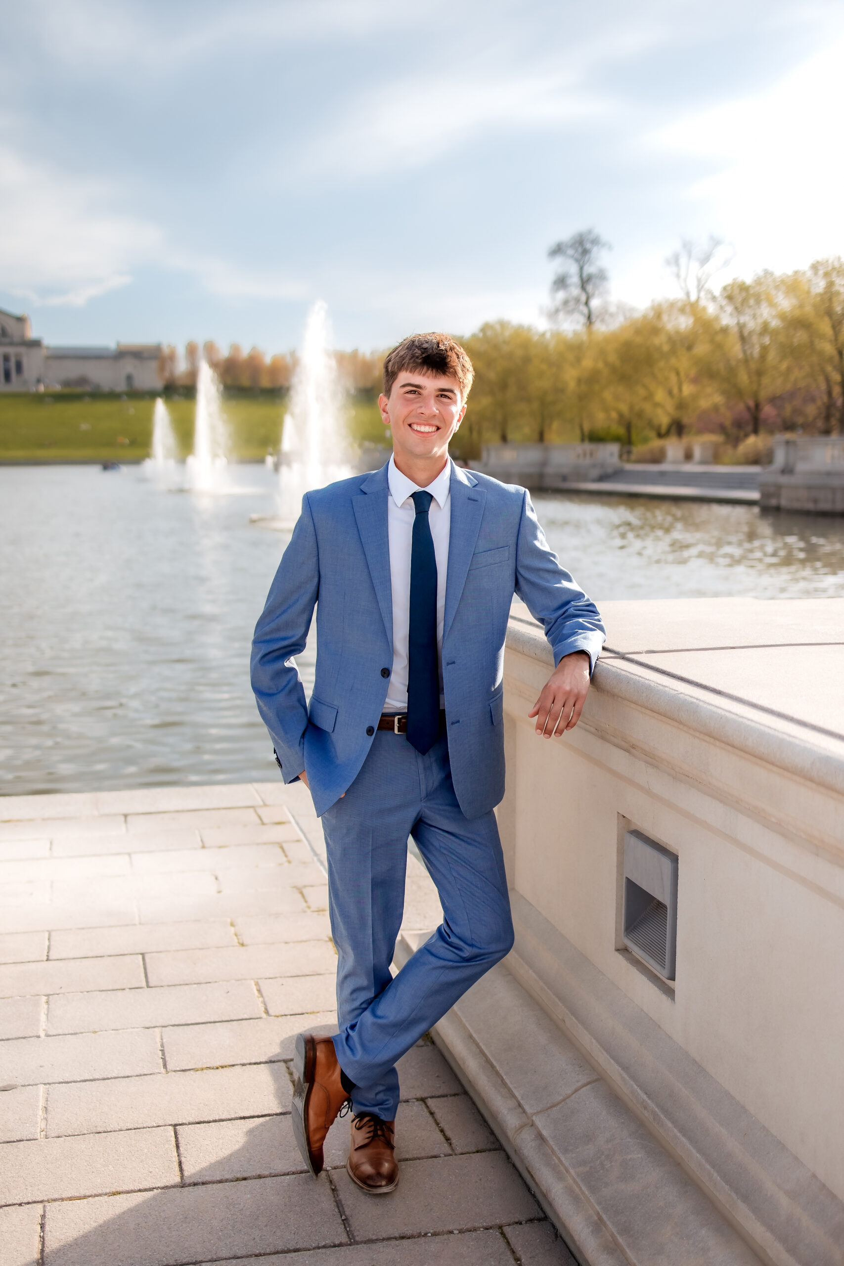 A young man is dressed in a blue suit and leaning against a stone wall, there is a pond behind him. He knows what to wear to your senior photos in St. Louis.