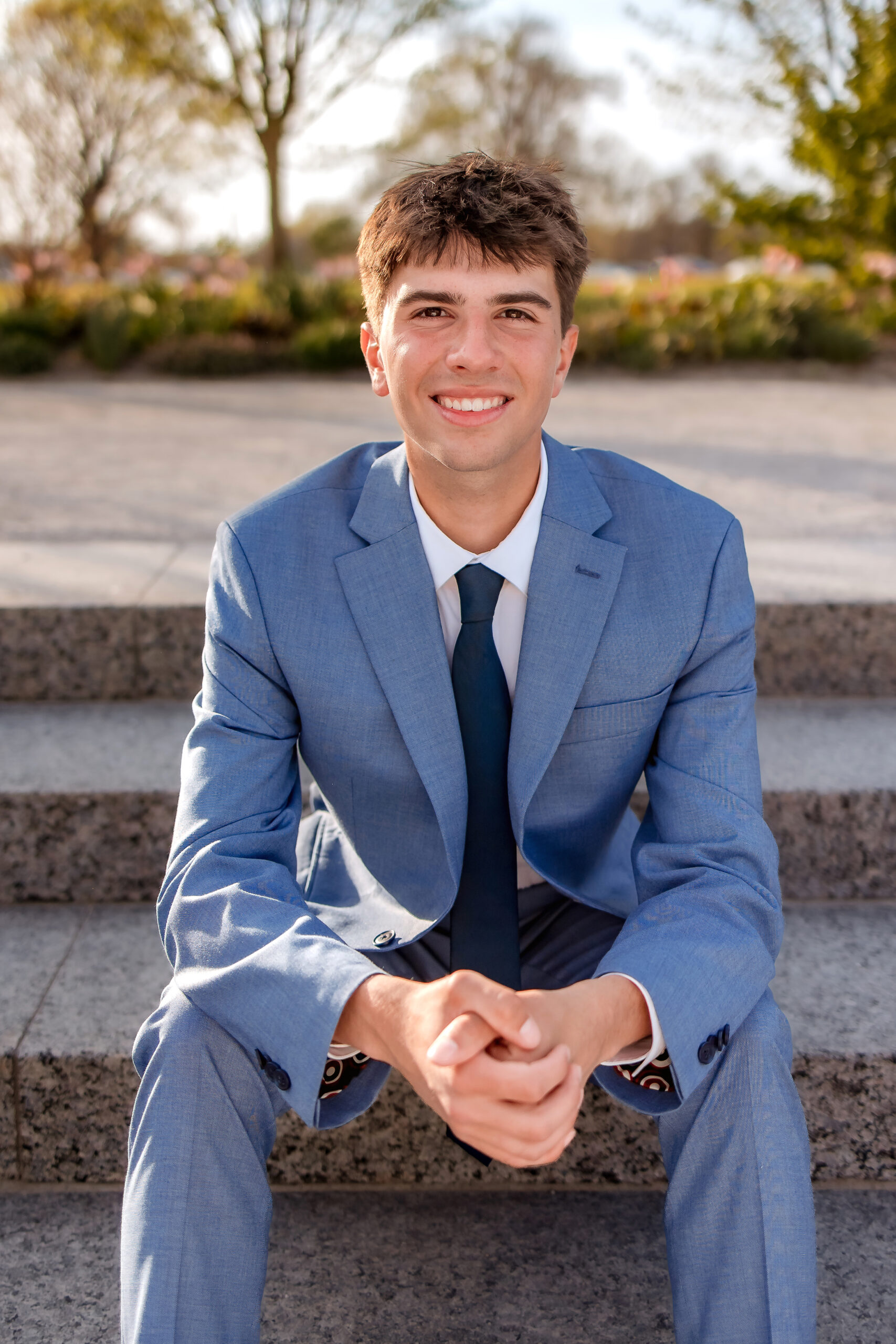 A young man dressed in a suit is sitting on some steps outside at the Grand Basin in St. Louis. His arms rest on his knees and his hands are closed together, he is smiling.