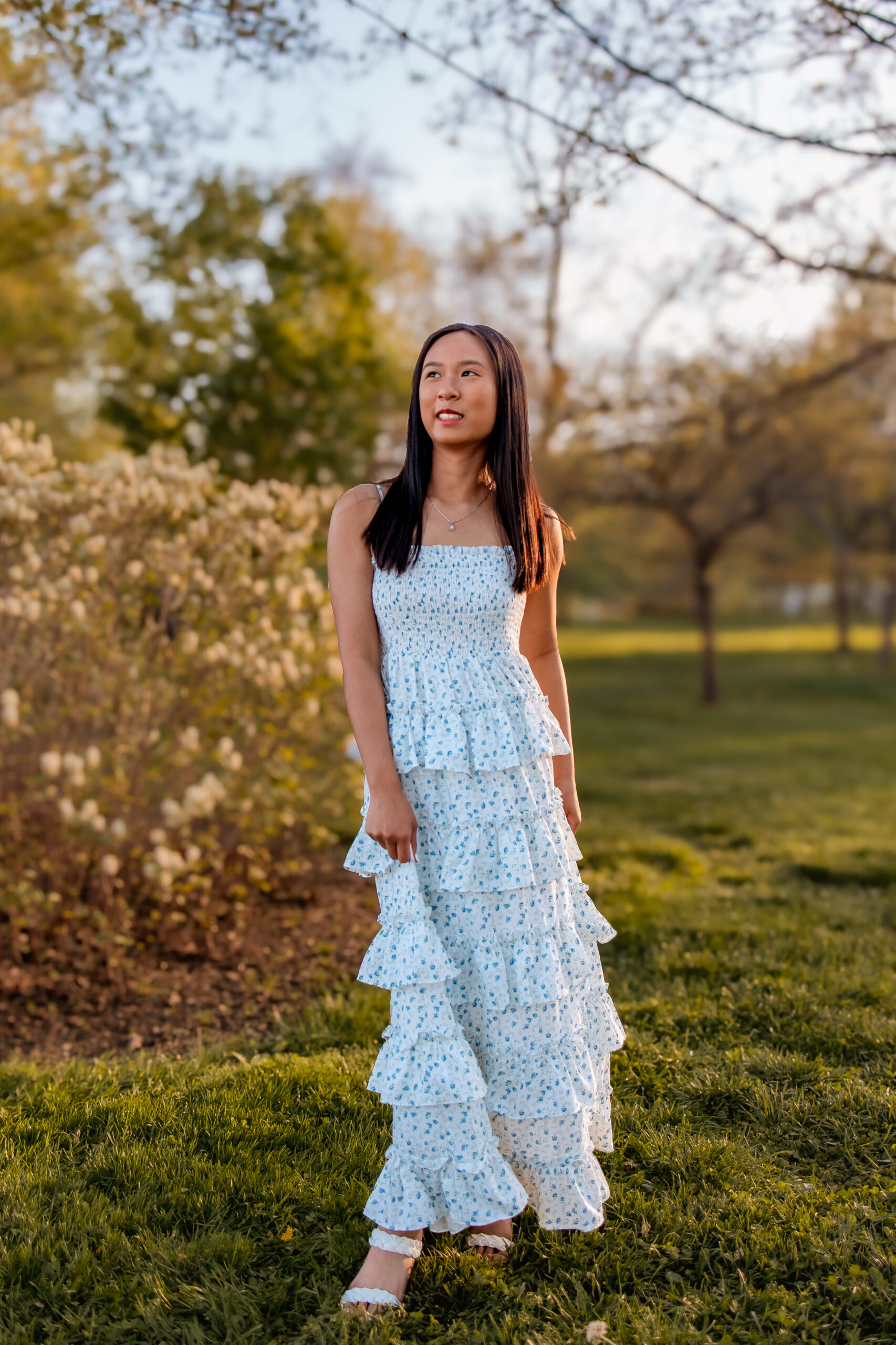A girl is dressed in a long ruffled dress and walking through the park looking off to the right at sunset.