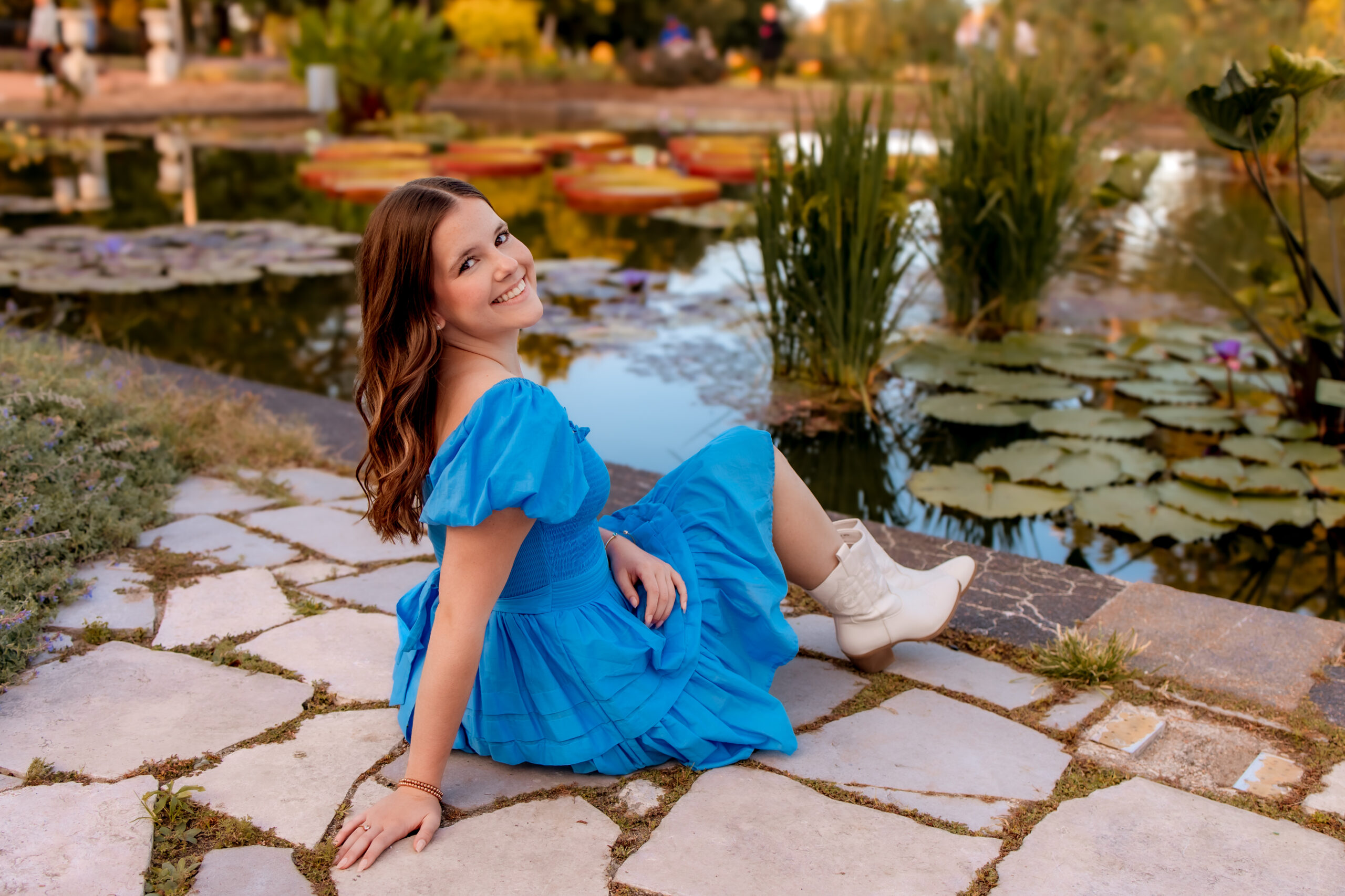 A girl is wearing a blue dress and sitting on a stone path in front of a pond with Lilly pads. There is a warm glow from sunset.