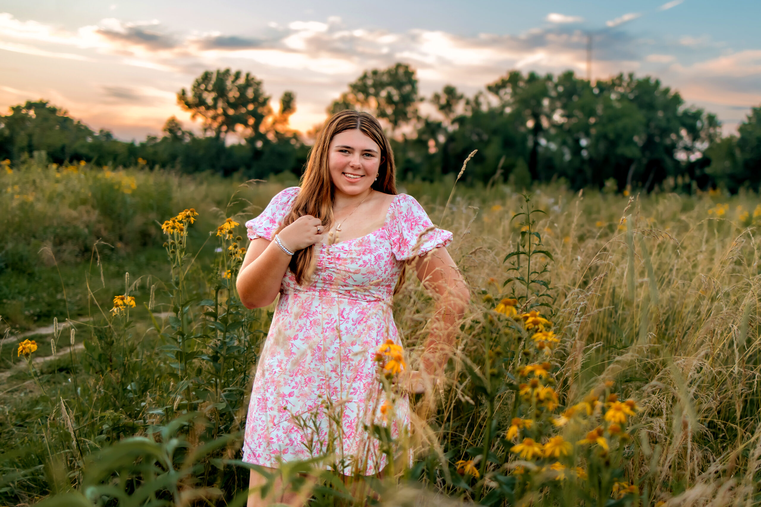 A girl is dressed in a pink and white floral dress. She is in a filed with wildflowers and left hand on her hip with right hand holder her necklace. she is smiling and there is a dynamic sunset with beautiful colors in the background.