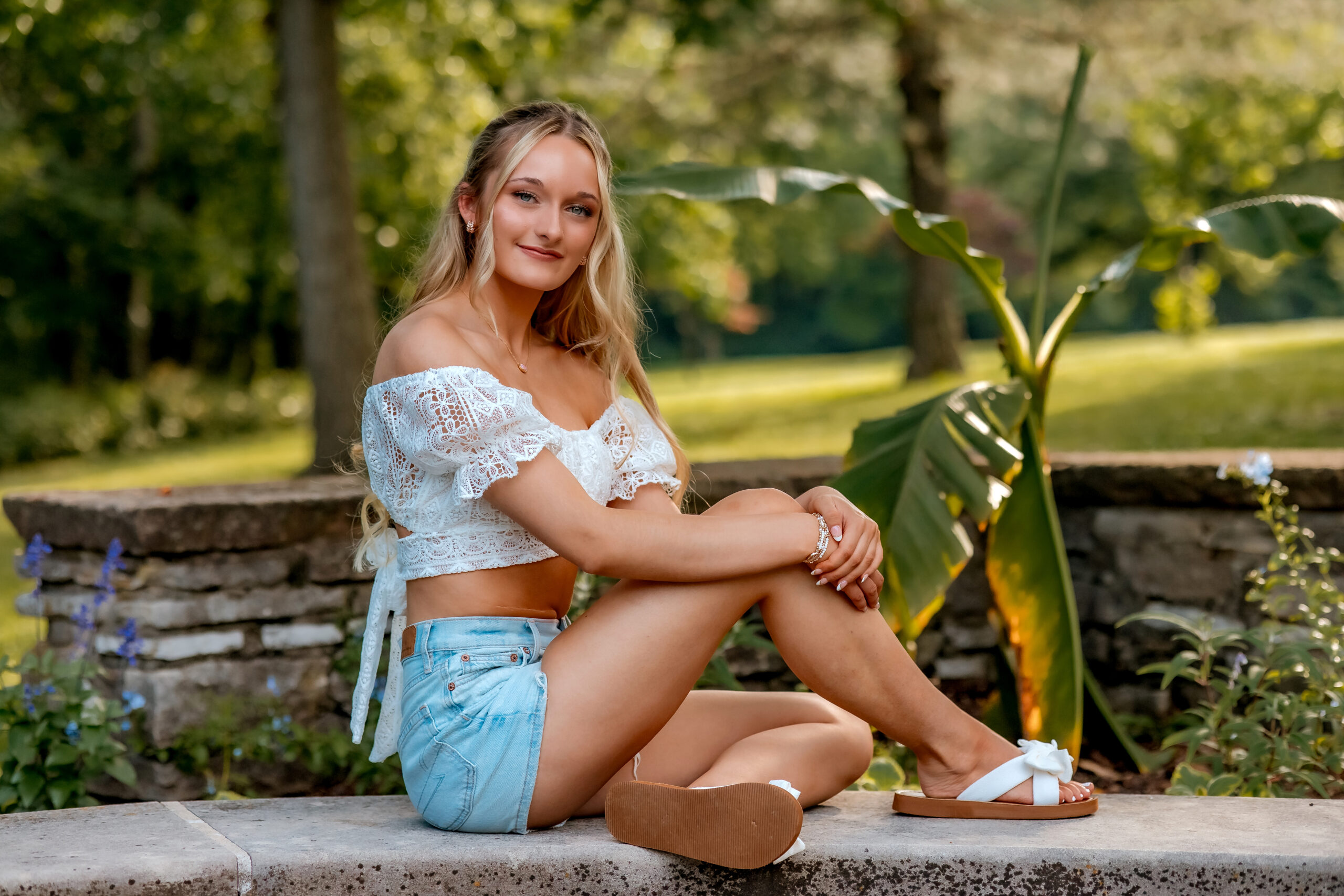 A girl is sitting on a concrete bench surrounded by greenery and banana trees and posing for her senior photos.