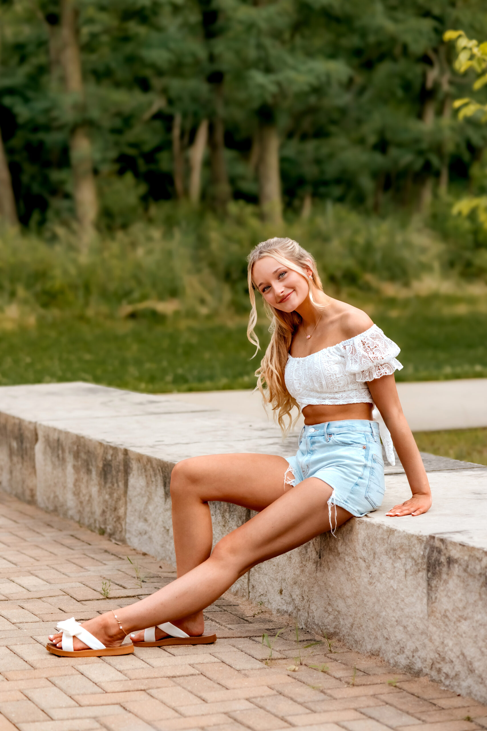 a girl is posing for her senior photos at Cliff Cave Park. She is sitting with her left leg stretched out and her right leg bent, her arms are grabbing the concrete she is stooped on and her back is tall with her head slightly leaning forward. Senior Photographs in St. Louis with Kaitlynn at Cliff Cave Park.