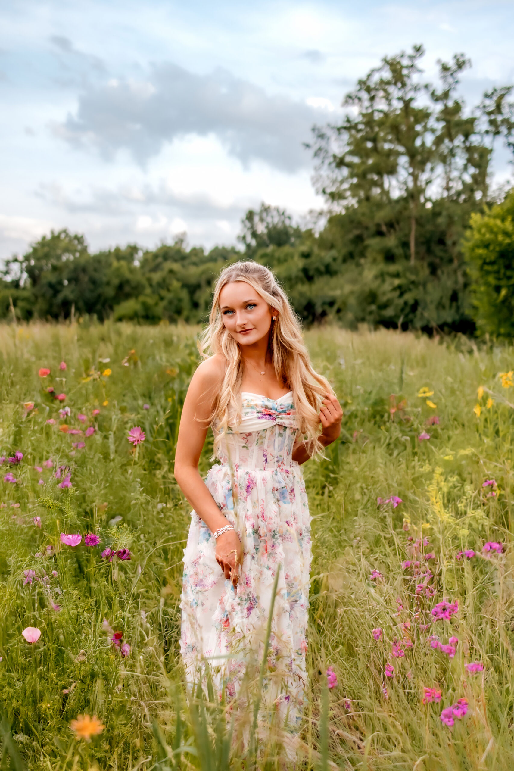A beautiful young girl with long wavy blonde hair is standing in the field of wildflowers at Cliff Cave park. Senior Photographs in St. Louis with Kaitlynn at Cliff Cave Park. She is holding the bottom of her hair with her left hand and the right hand is holding her long floral dress.