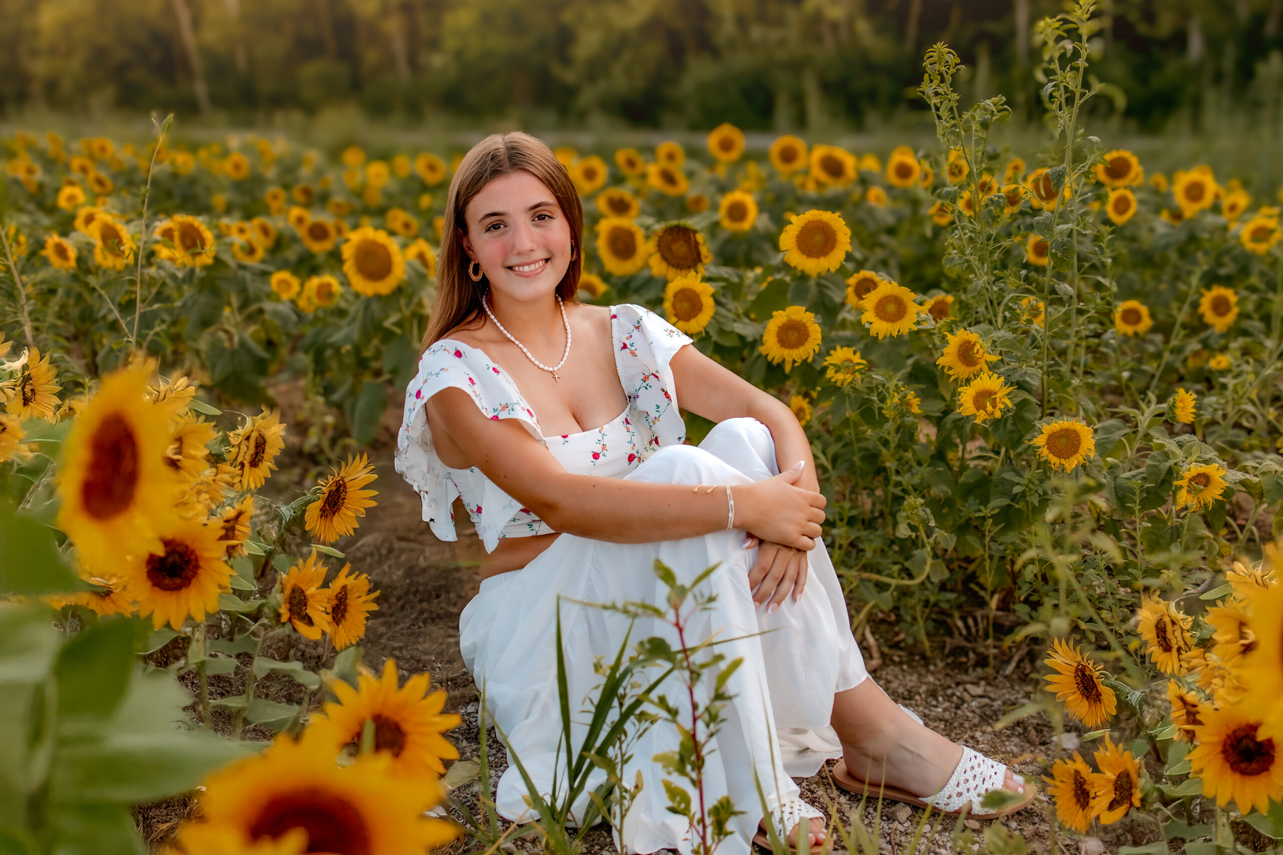 A brunette girl is sitting in a field of sunflowers. Her arms are wrapped around her knees. There is a warm glow from sunset.