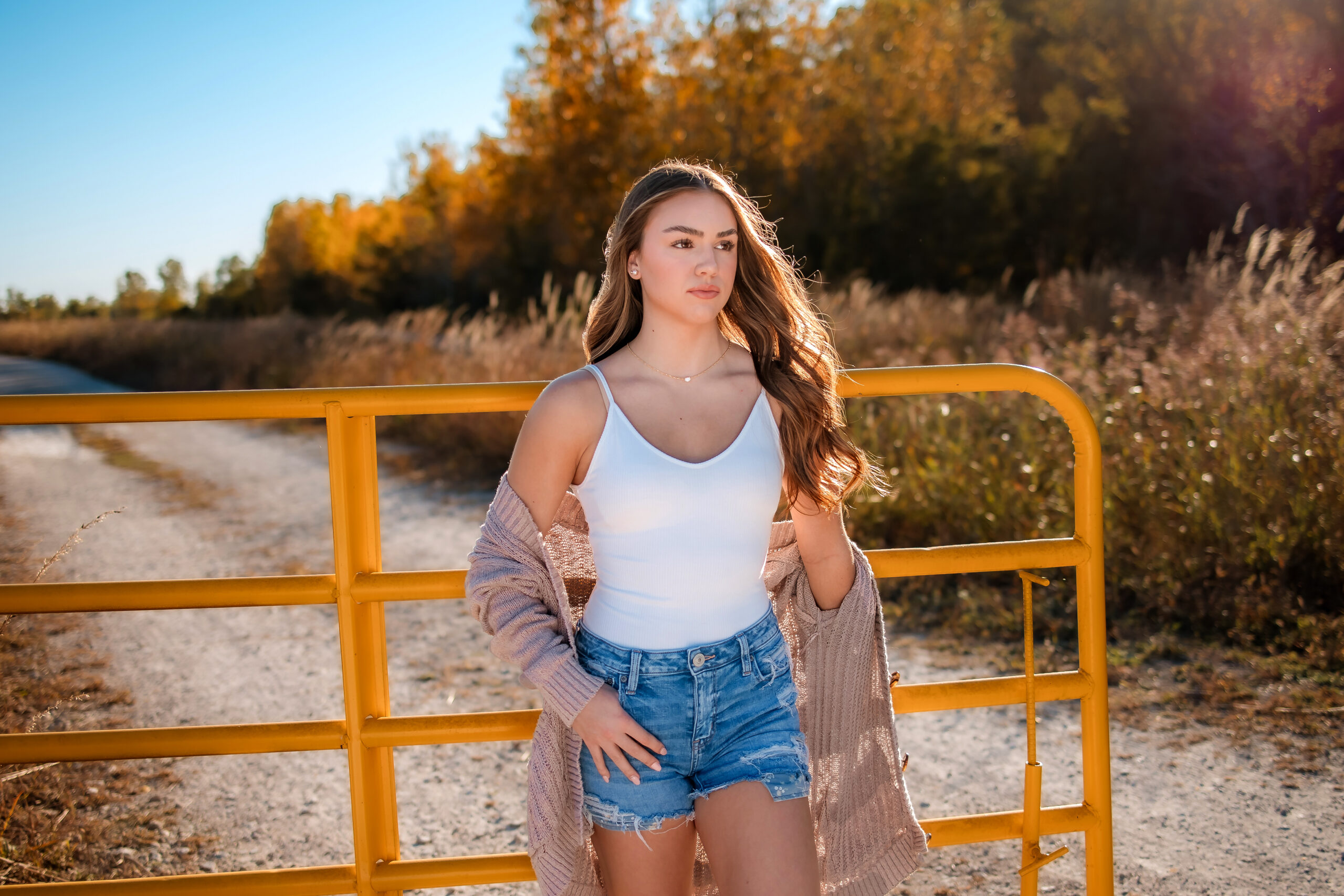 A girl with long brown hair is leaning against a yellow country gate with dirt road behind it. She look very serious and is looking to the left. She has on denim shorts, a white tank top and has a pink fuzzy cardigan draped off her shoulders. The sun is shining and backlighting the subject.