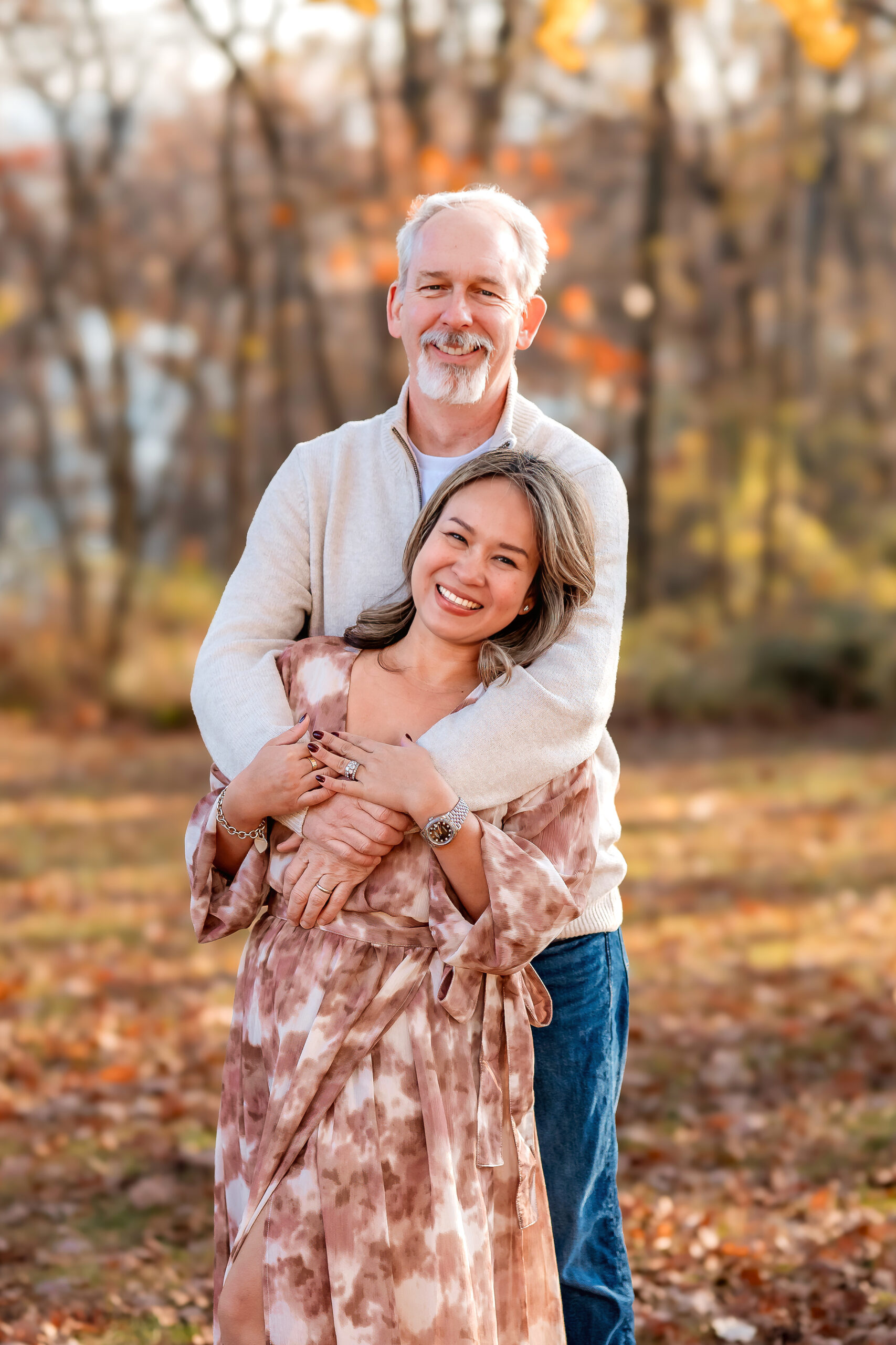 an older couple is holding each other and while they both laugh and smile towards the camera. They are in a park with fall leaves fallen and still on the trees.