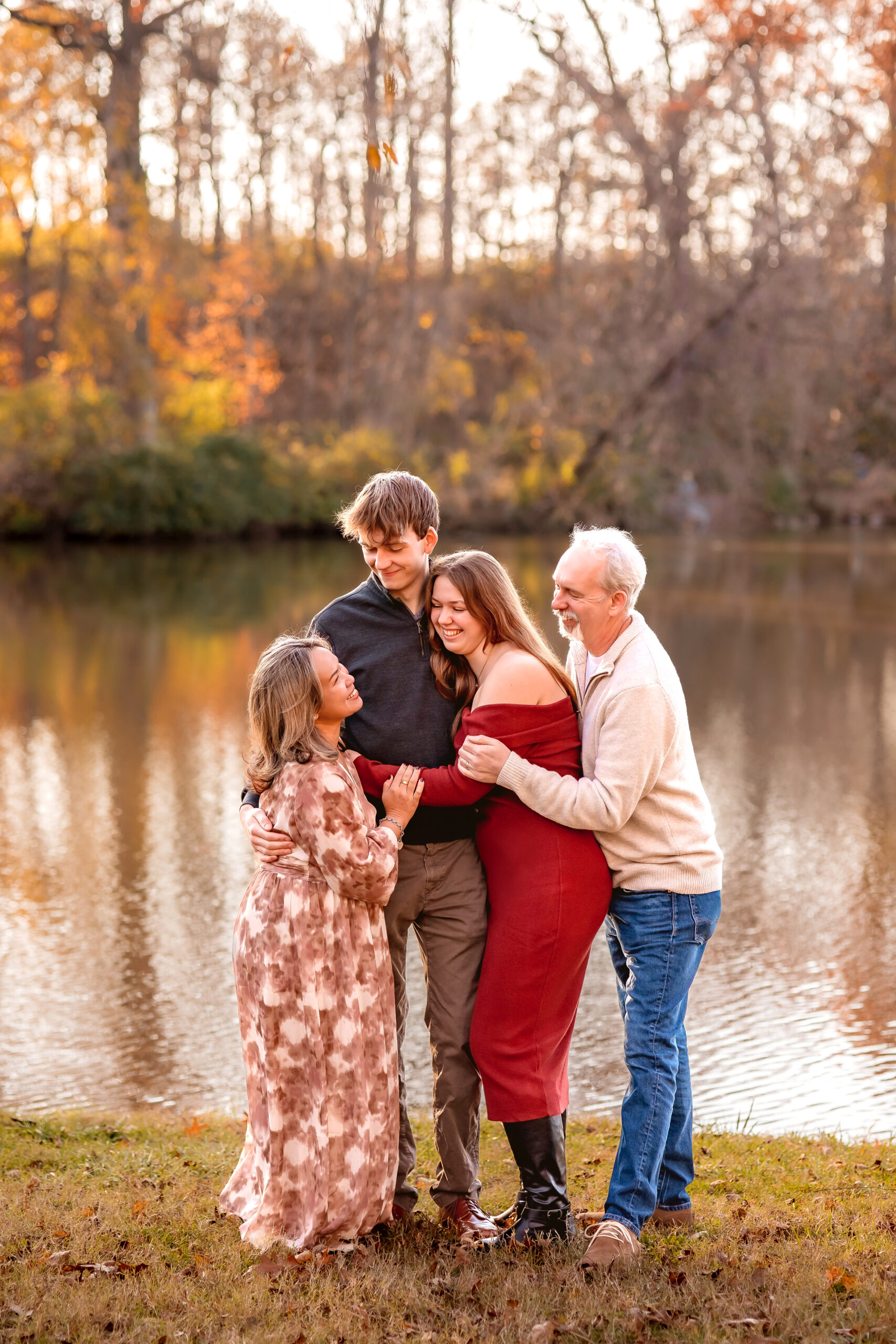 a beautiful landscape of a family of 4 hugging and laughing with the morning sunshine glow on the colorful fall leaves and the reflections bouncing off the pond in the background.