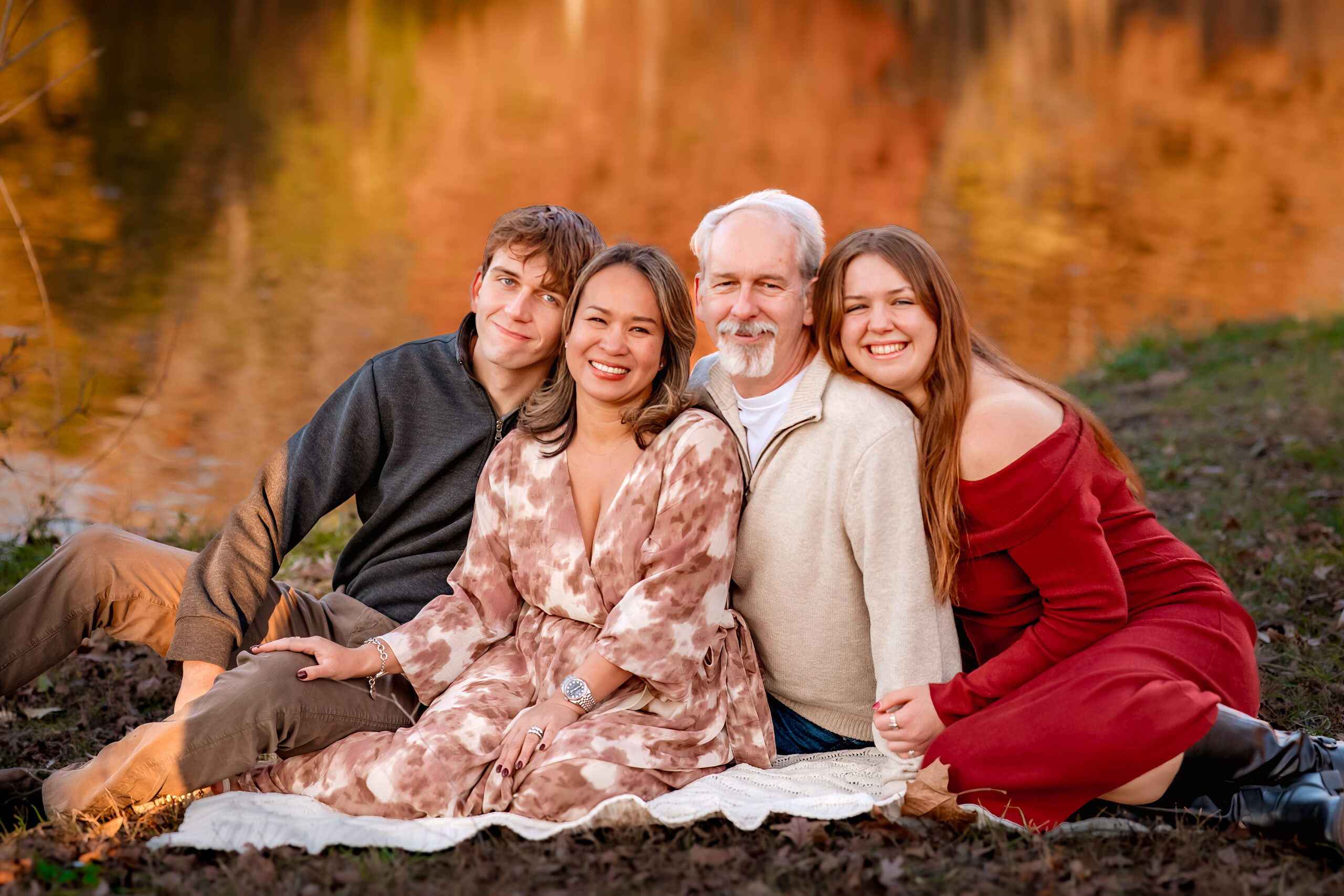 a family of four is snuggled on a blanket in the park sitting near a pond in fall with the morning sunshine glow.